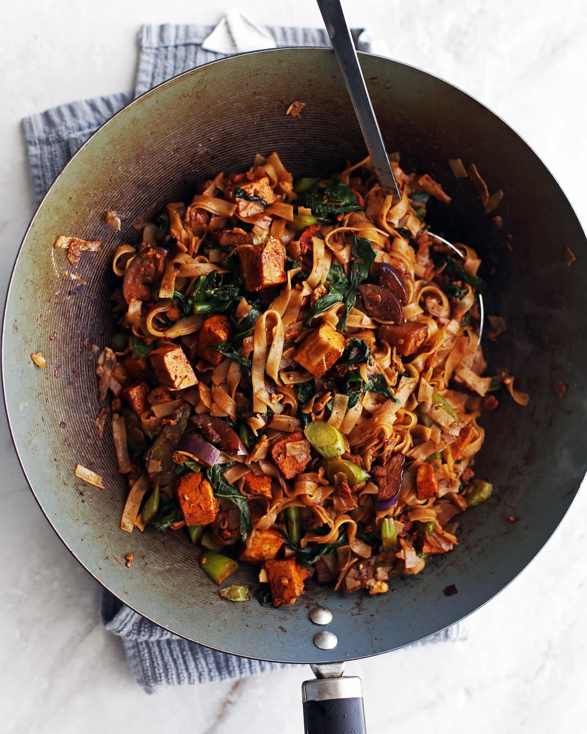 rice noodles, eggplant, tofu, and Thai basil all together in a wok getting tossed with a pair of tongs