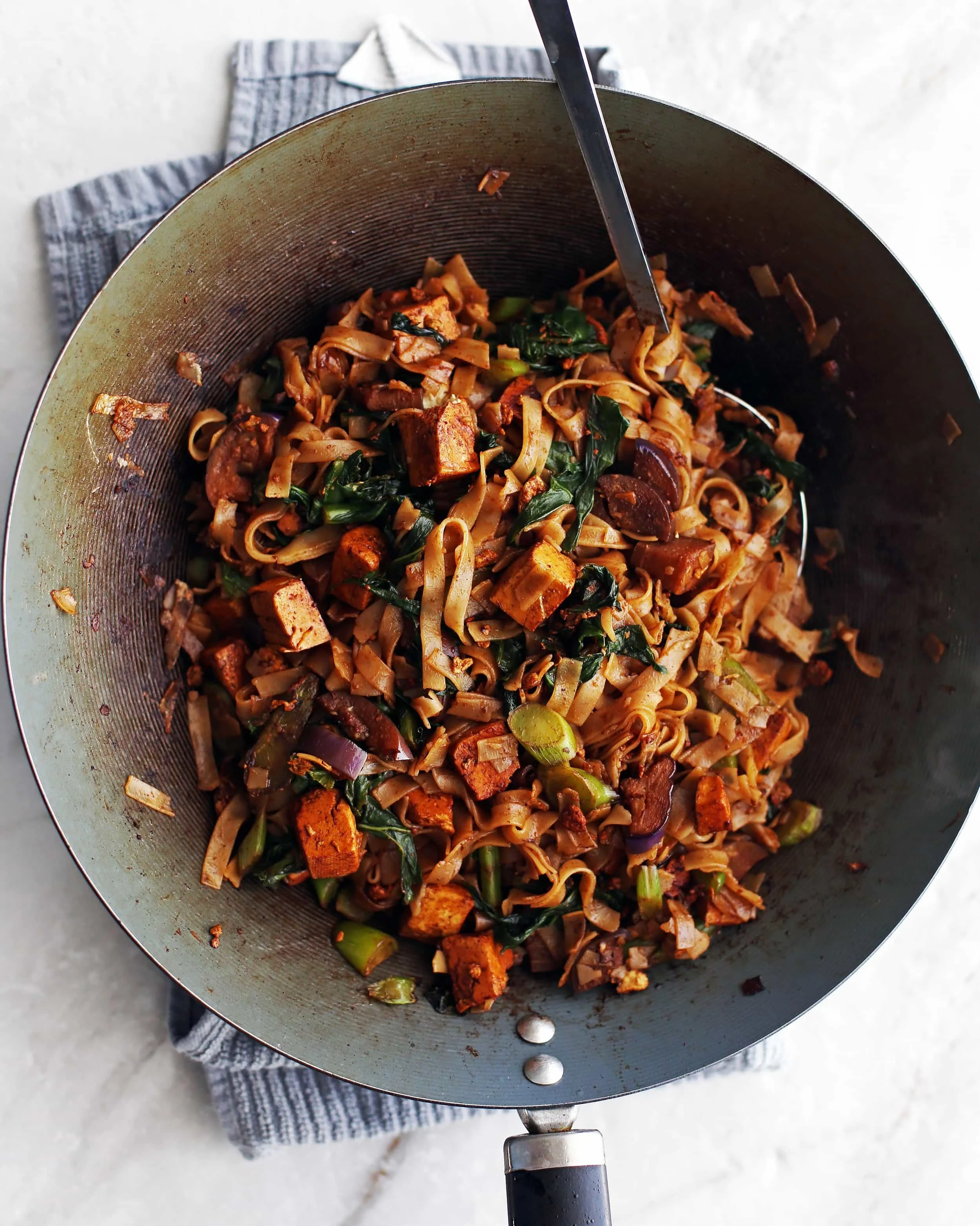 rice noodles, eggplant, tofu, and Thai basil all together in a wok getting tossed with a pair of tongs