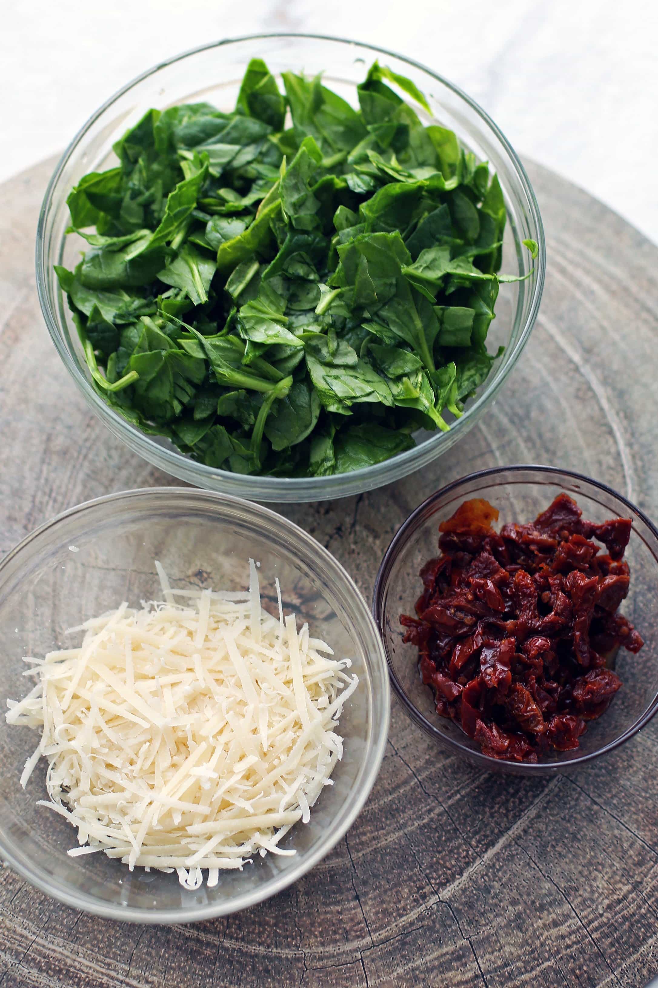 Three glass bowls containing freshly grated parmesan cheese, chopped sun-dried tomatoes, and chopped baby spinach.