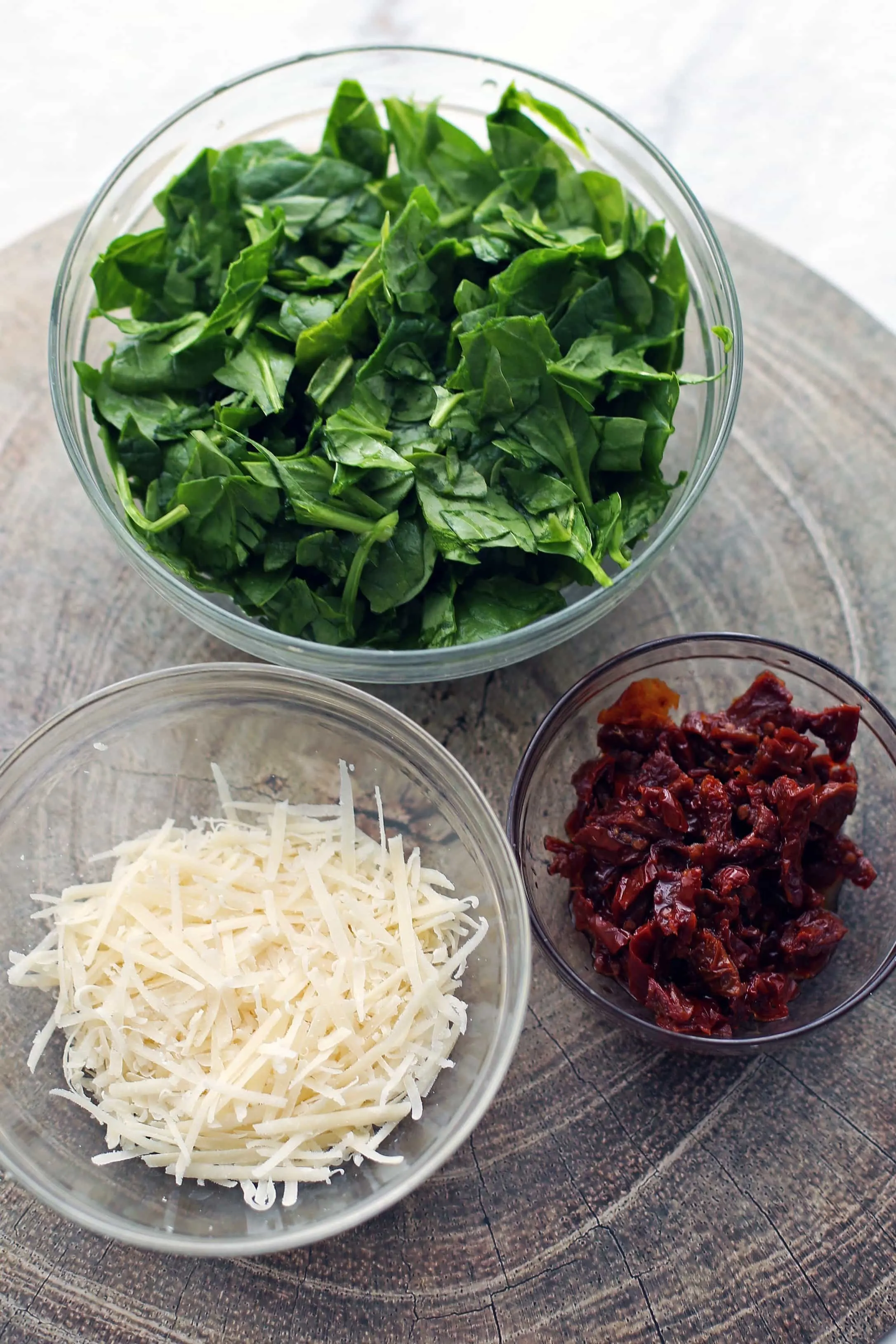 Three glass bowls containing freshly grated parmesan cheese, chopped sun-dried tomatoes, and chopped baby spinach.