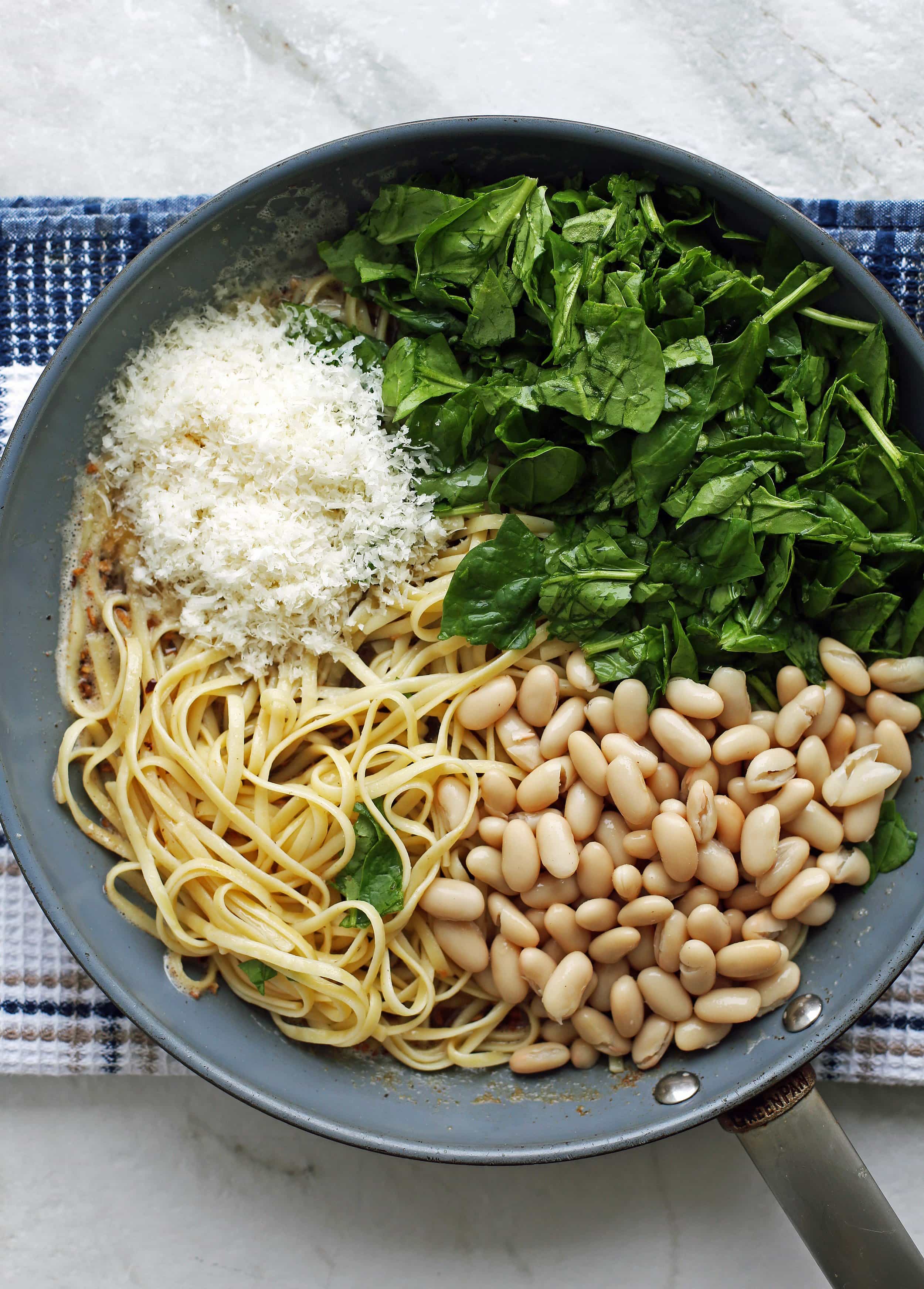 Pasta, white beans, spinach, and parmesan cheese placed on top of a brown butter sauce that’s in a large skillet.
