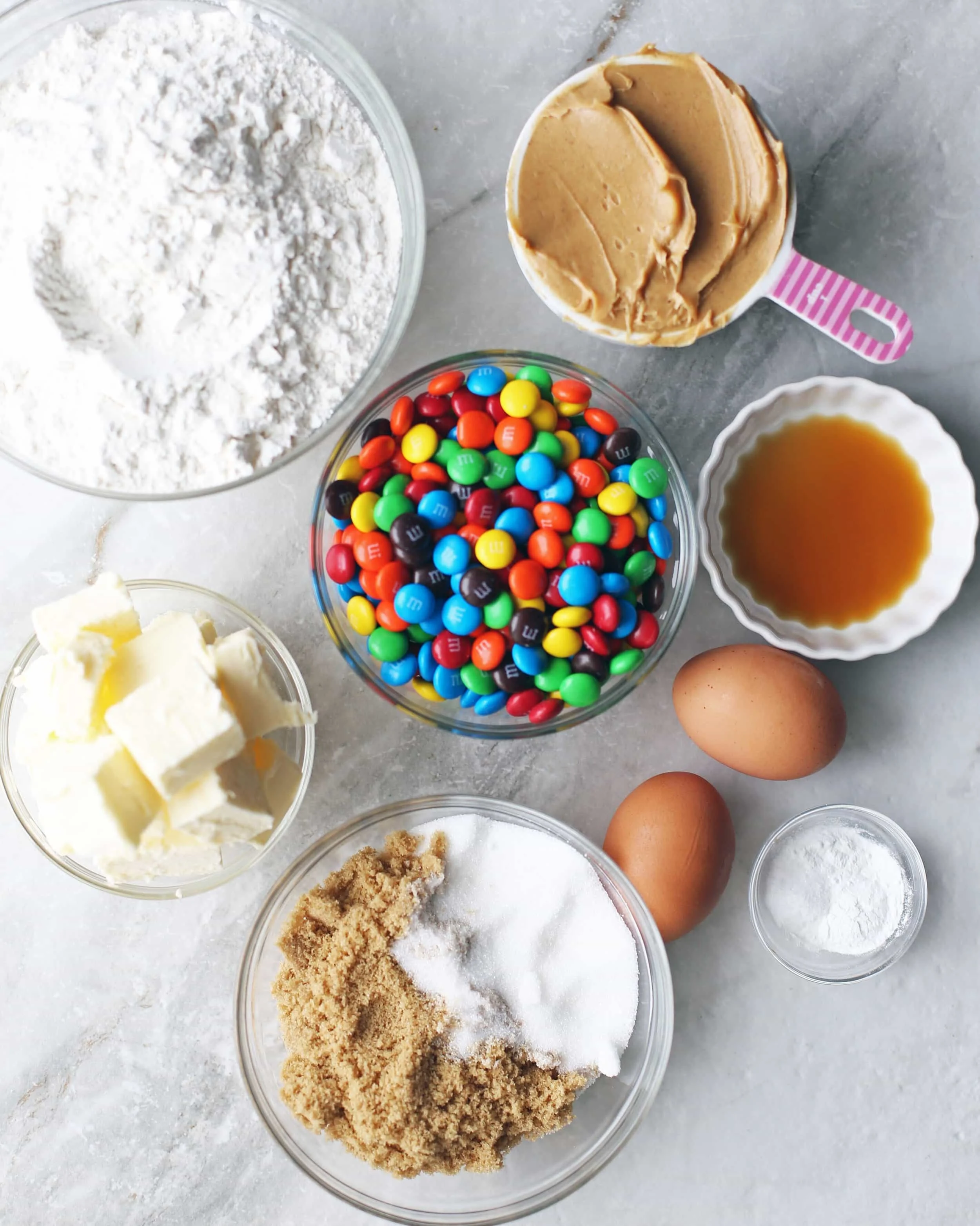 Overhead shot of glass bowls filled with white and brown sugar, butter, flour, peanut butter, vanilla extract, M&M's, eggs, salt, and baking powder.