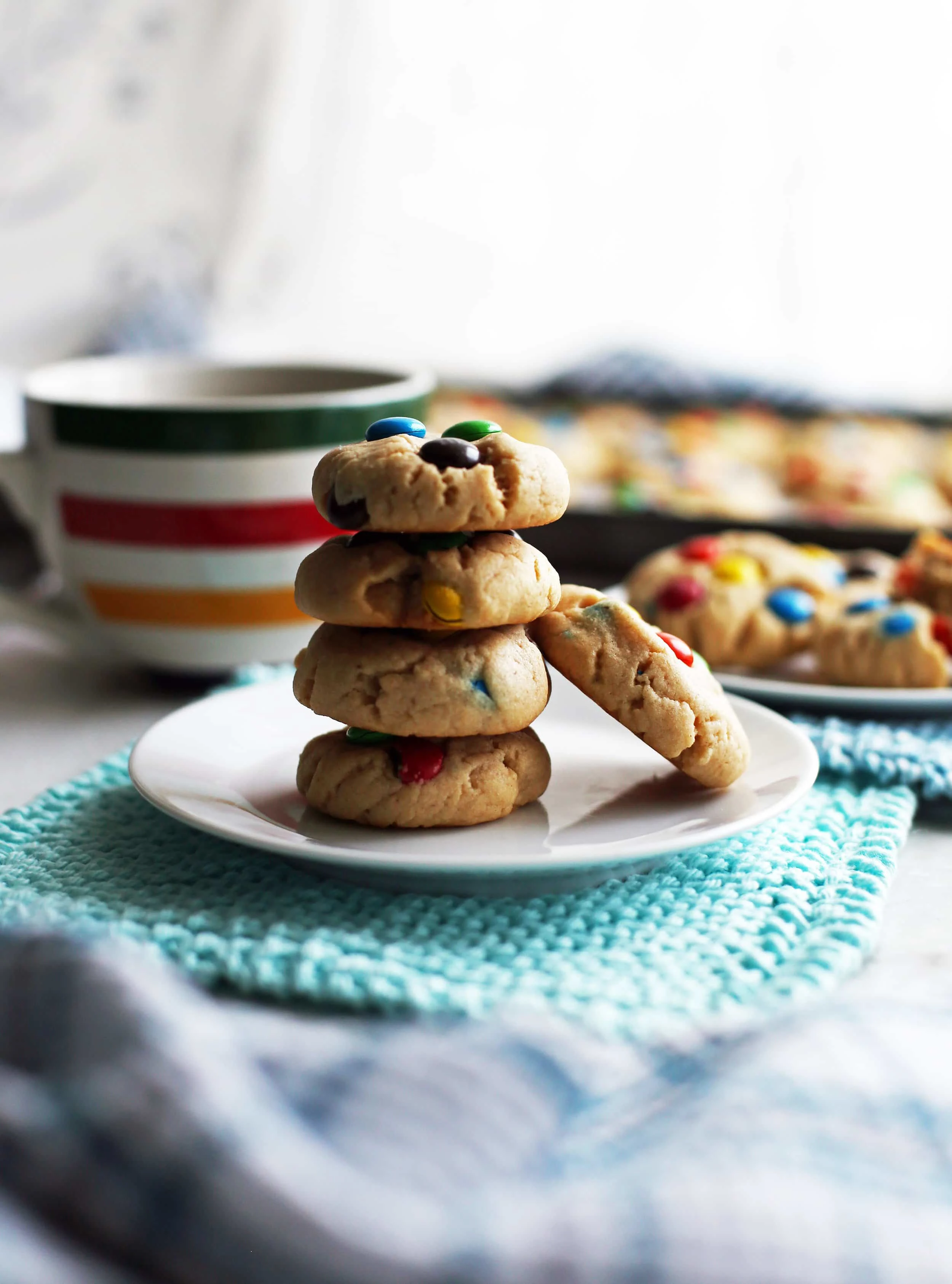 Four stacked peanut butter M&M's cookies on a white plate with more cookies in the background.