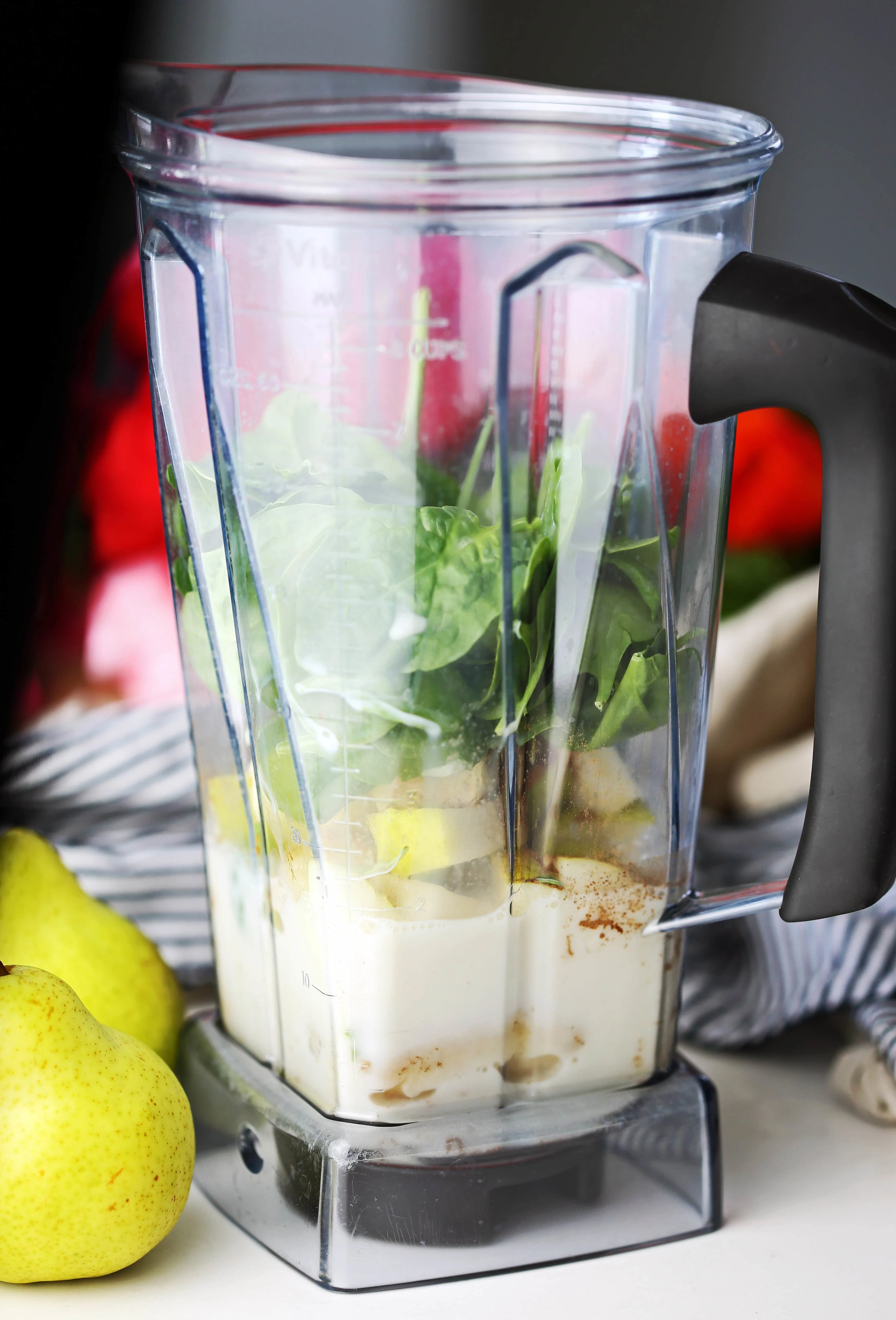 A blender container holding chopped pears, banana, spinach, ground cinnamon, almond milk, vanilla, and maple syrup.
