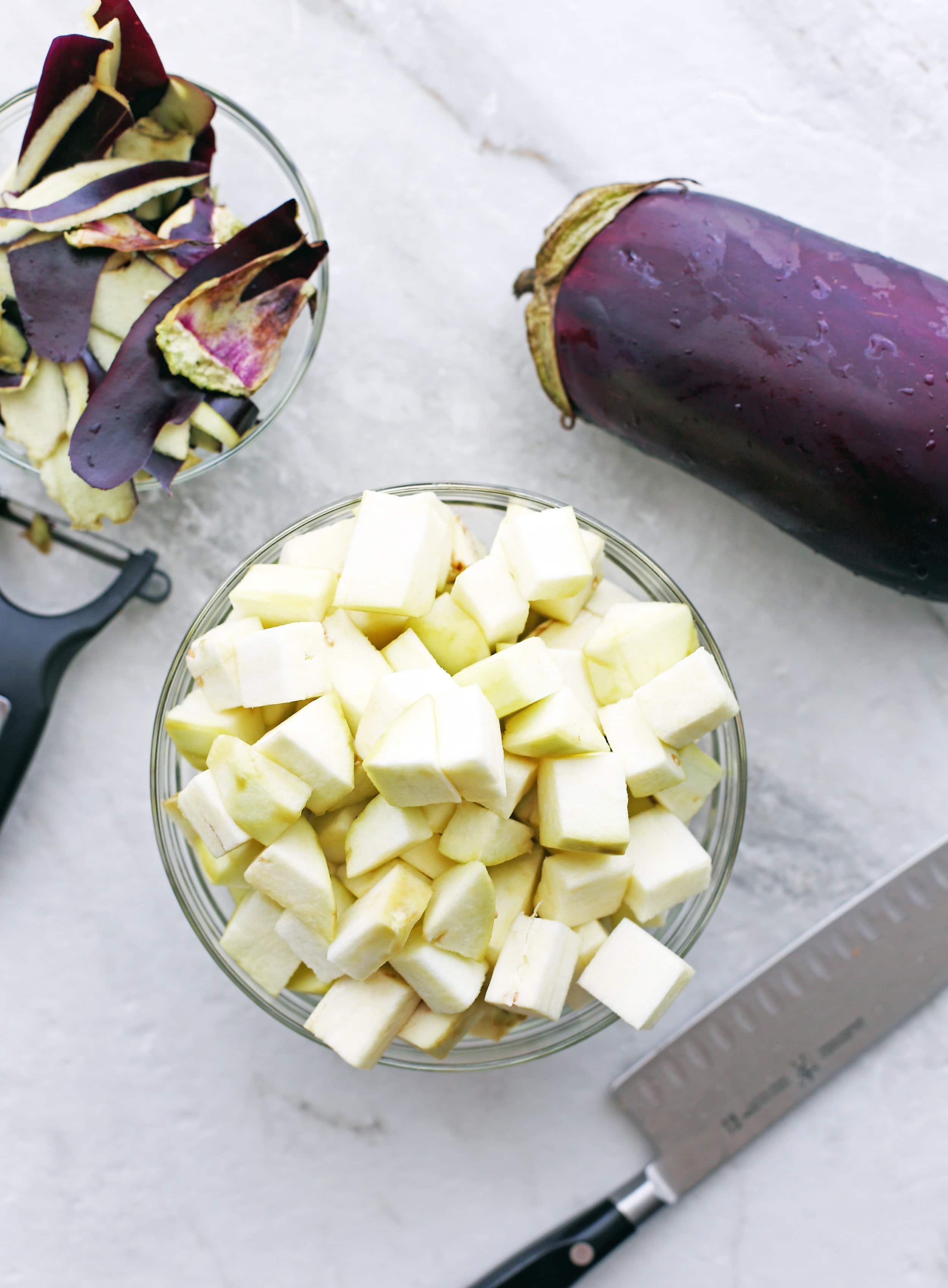 Peeled and chopped Italian eggplant in glass bowl with knife, another eggplant, and eggplant skin around the bowl.