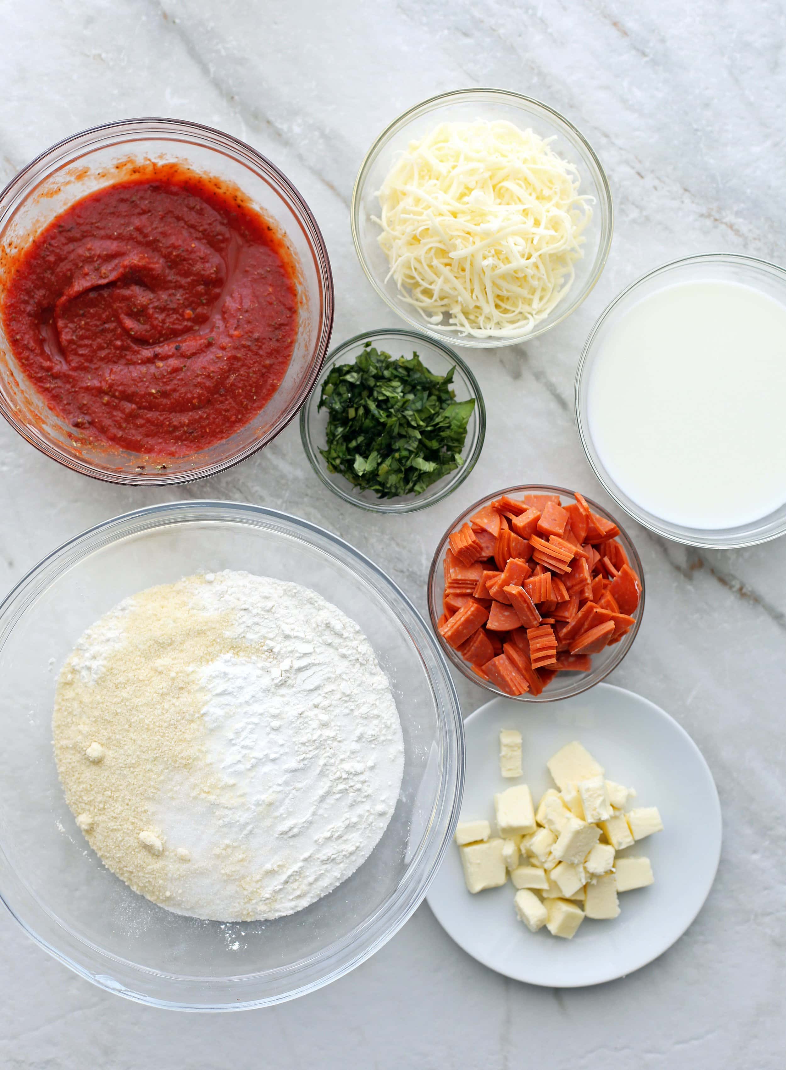 Bowls of pepperoni, cheese, fresh basil, pizza sauce, flour mixture, milk, and butter.