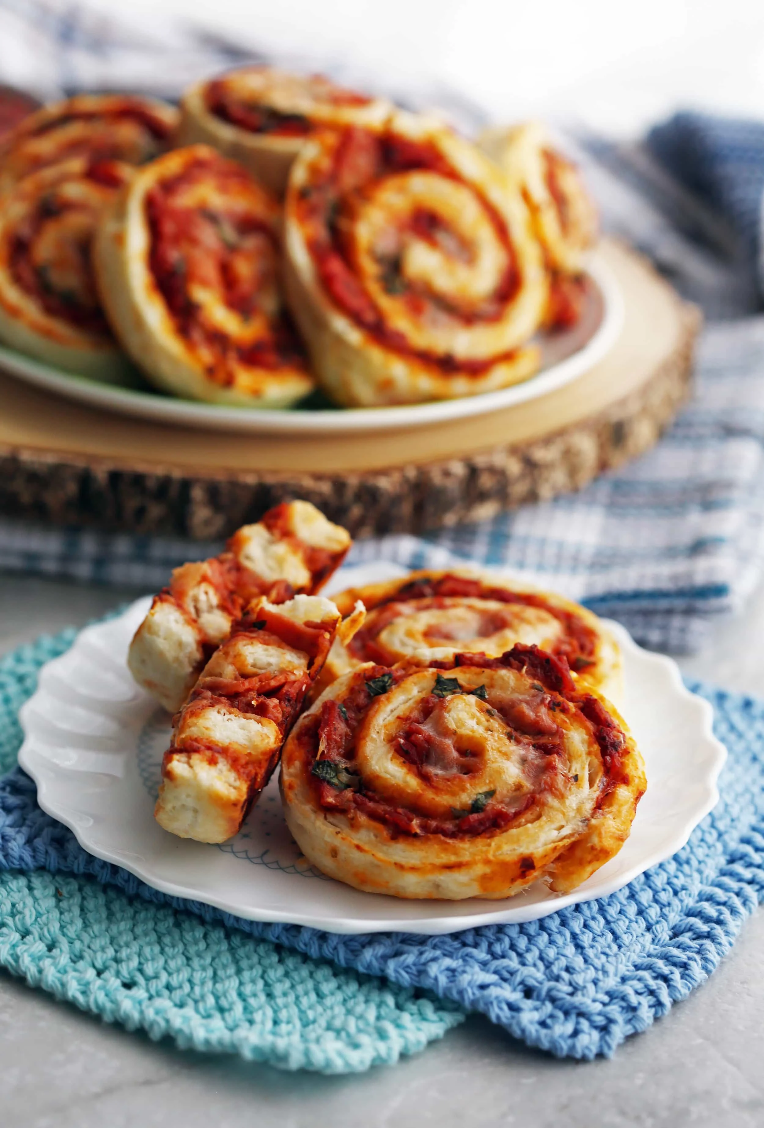 Three baked pepperoni cheese pizza rolls on a white plate with more pizza rolls piled on a plate in the background.