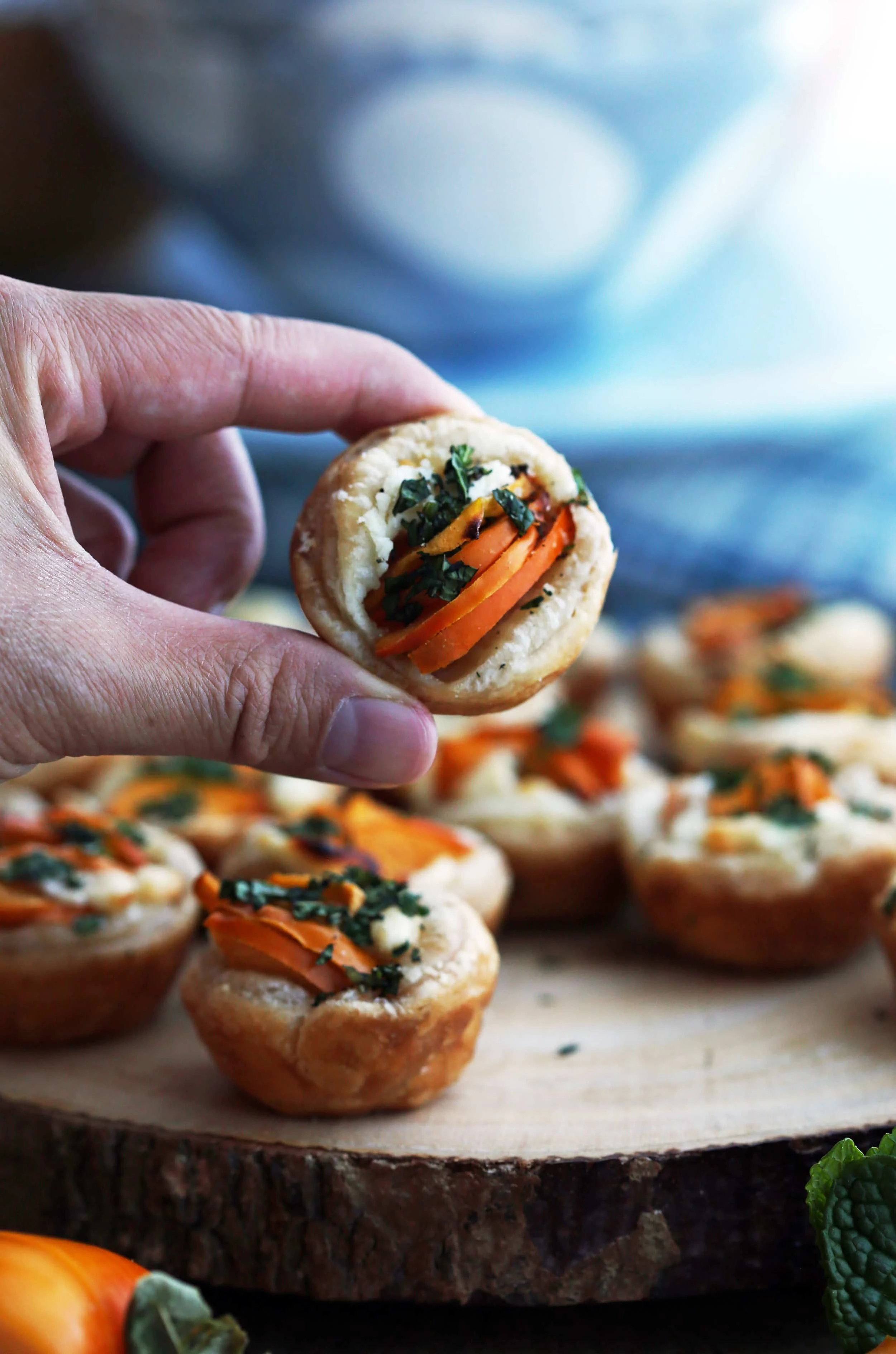 A hand holding a single Persimmon Goat Cheese Tartlet over a wooden platter full of more tartlets.