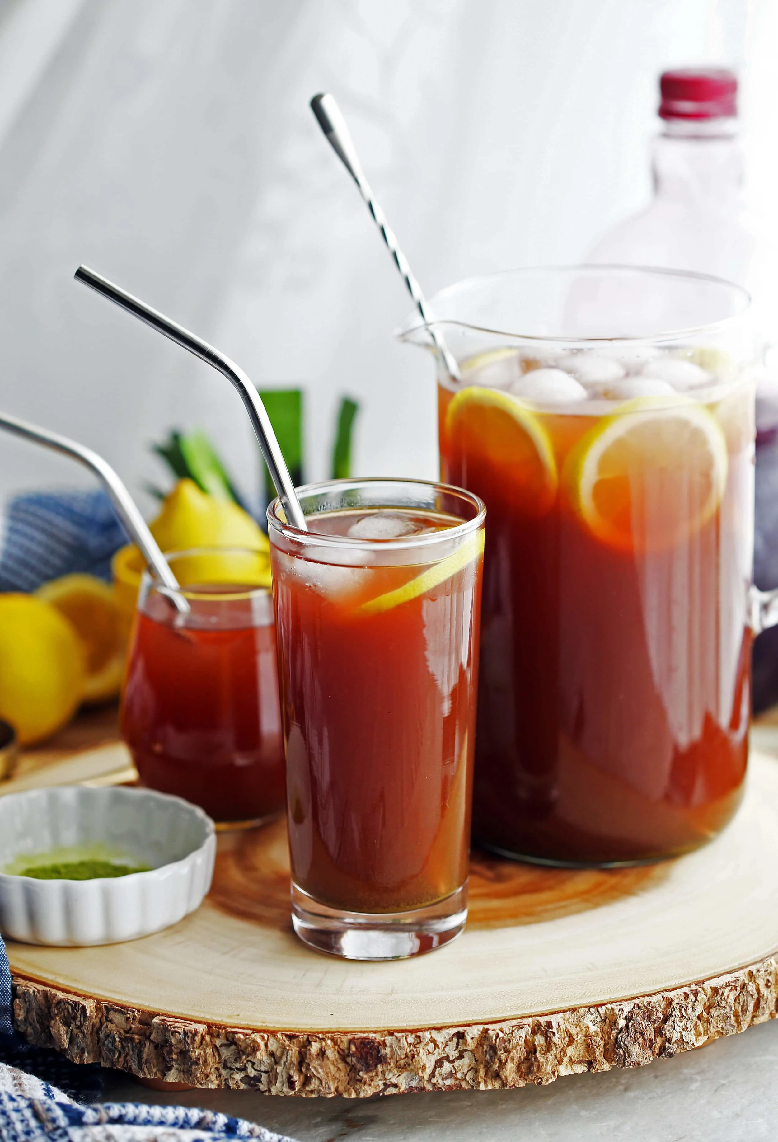 Pomegranate matcha iced tea with lemon slices and ice in two glasses and a glass pitcher.