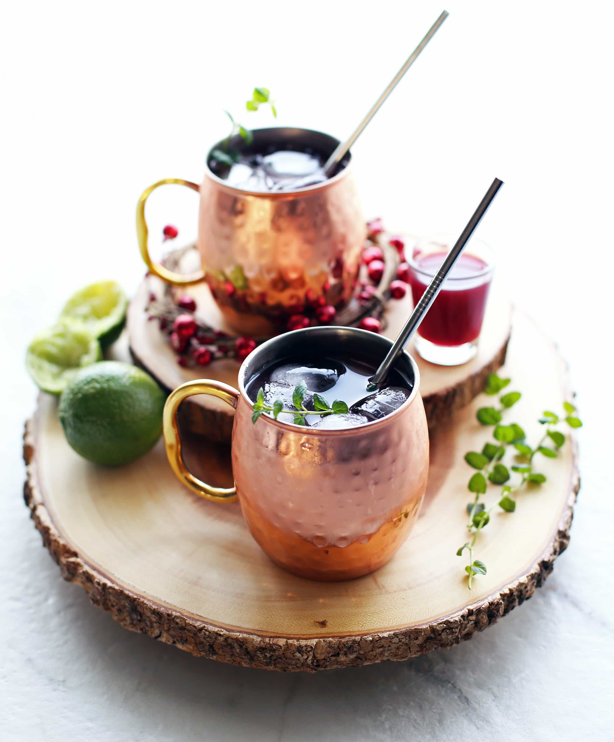 Two Pomegranate Honey Moscow Mule cocktails in copper mugs on two round wood slabs.