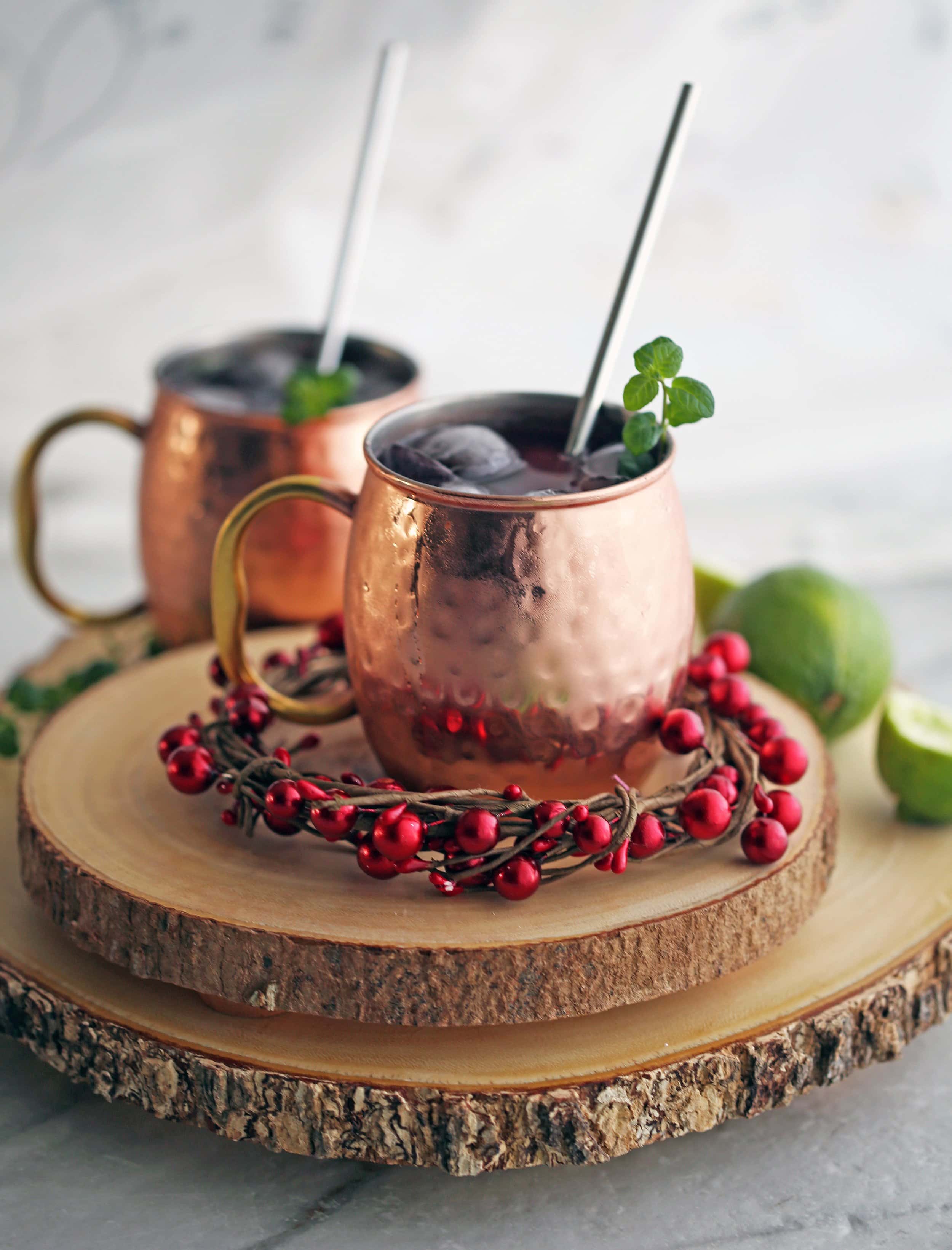 Two Pomegranate Honey Moscow Mule cocktails in copper mugs on wood slabs.