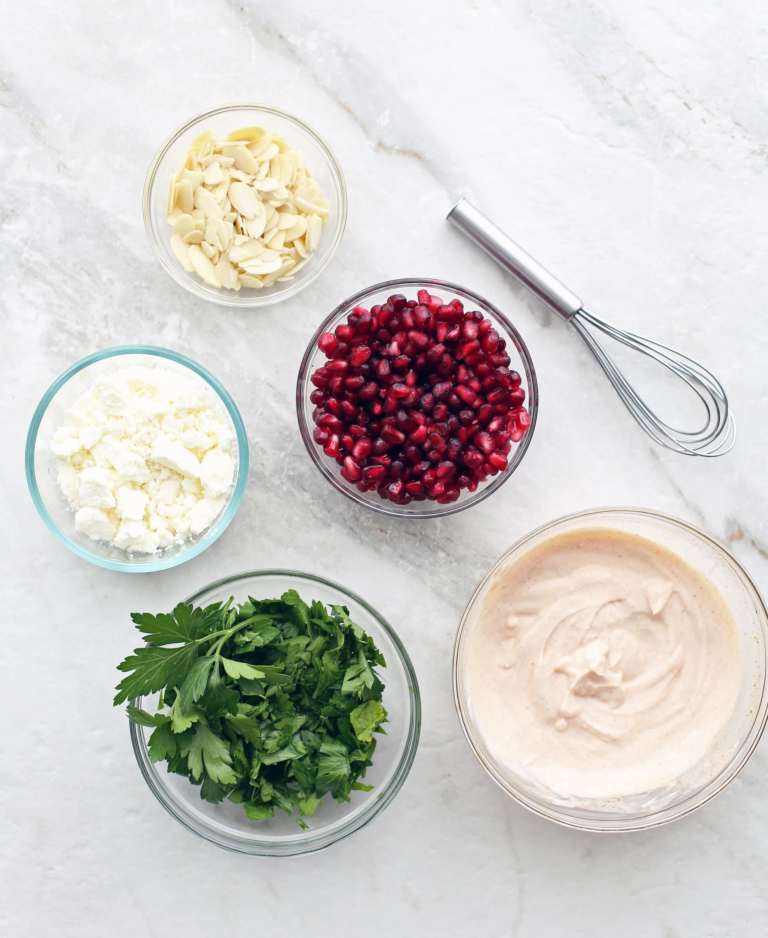 Bowls containing feta cheese, parsley, pomegranate arils, slivered almonds, and paprika yogurt dressing.