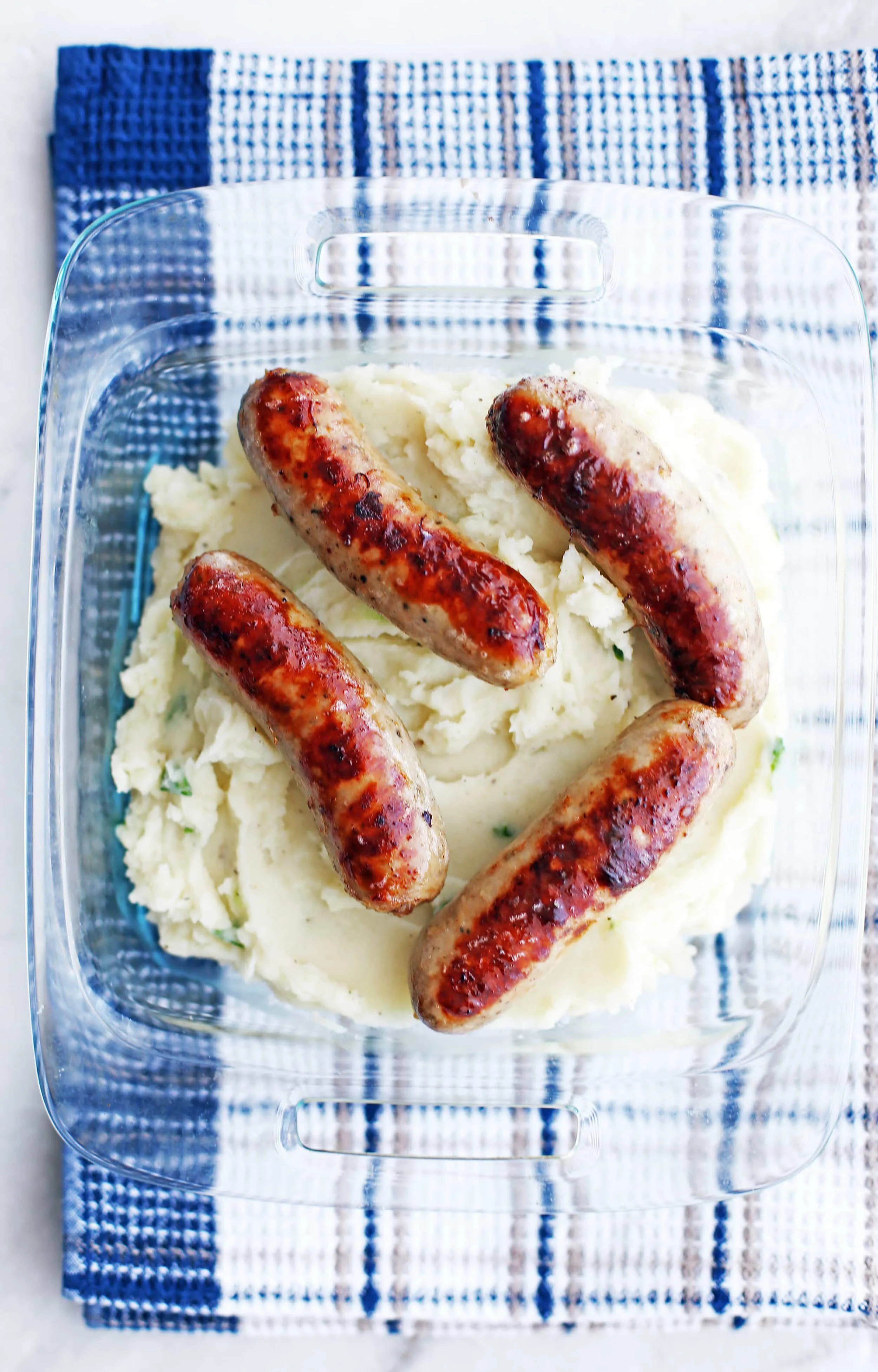 Creamy mashed potatoes with four cooked golden-brown pork sausages on top in a glass square dish.