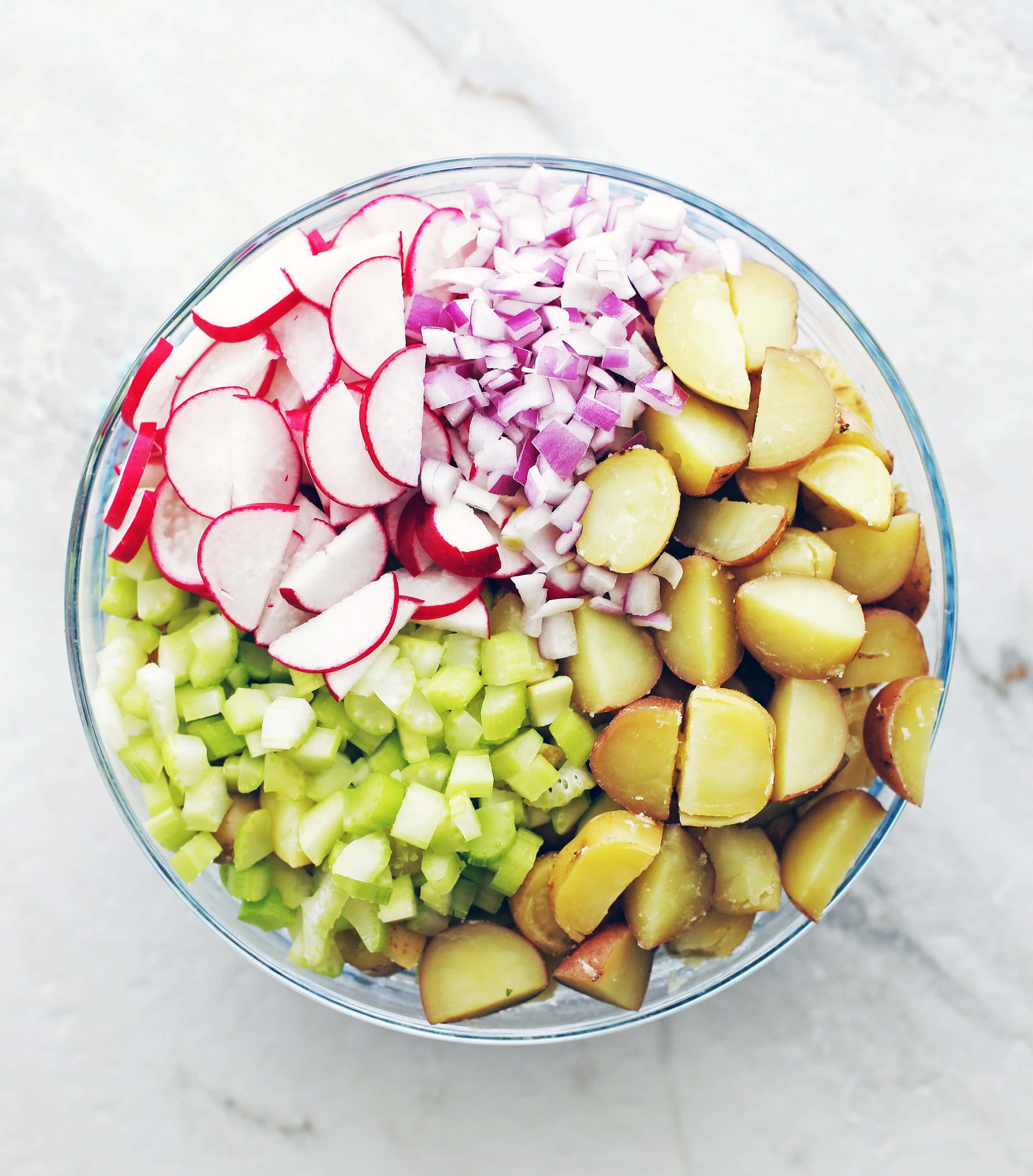 Boiled sliced potatoes, diced celery and red onions, and sliced radishes in a large glass bowl.