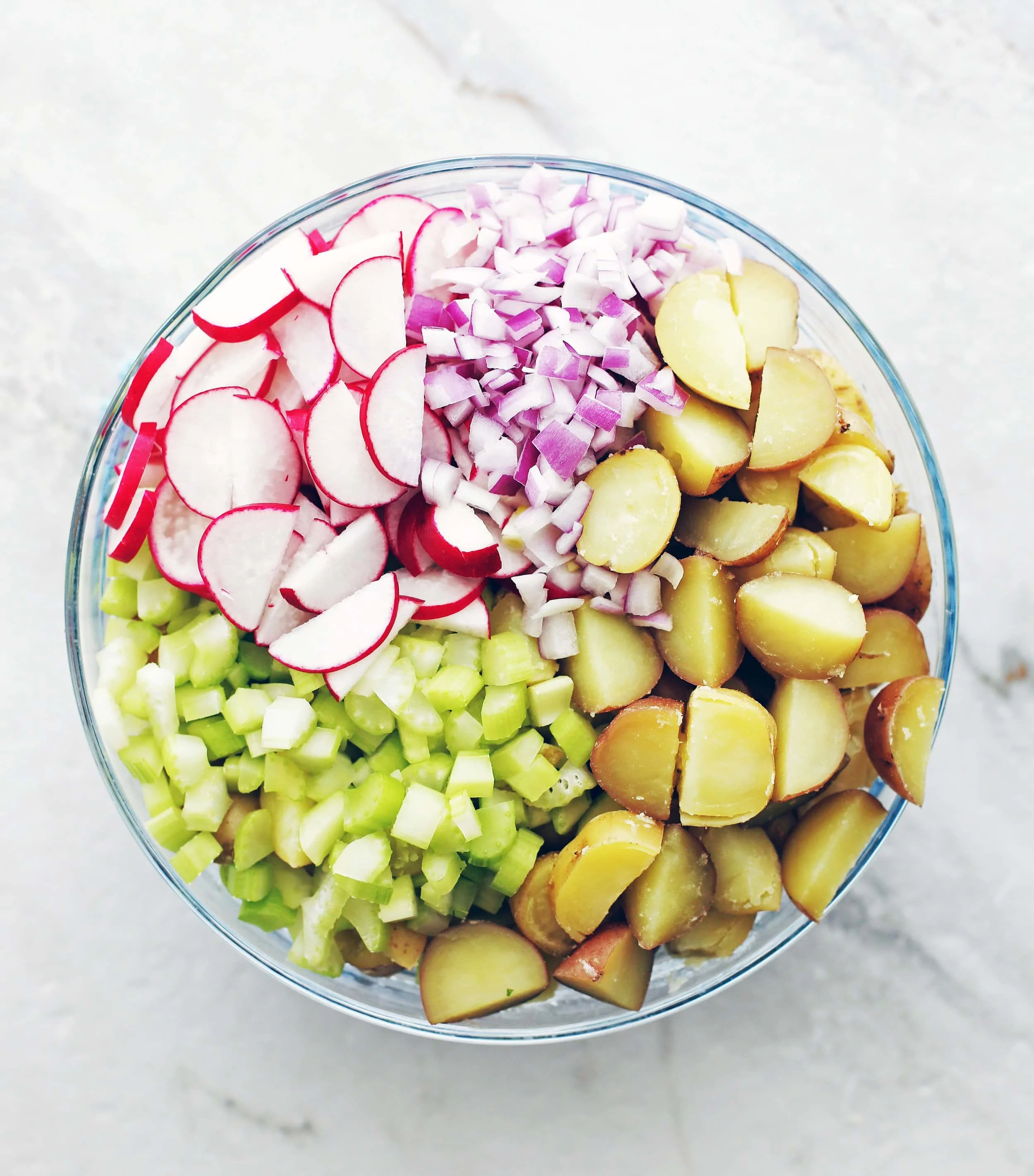Boiled sliced potatoes, diced celery and red onions, and sliced radishes in a large glass bowl.