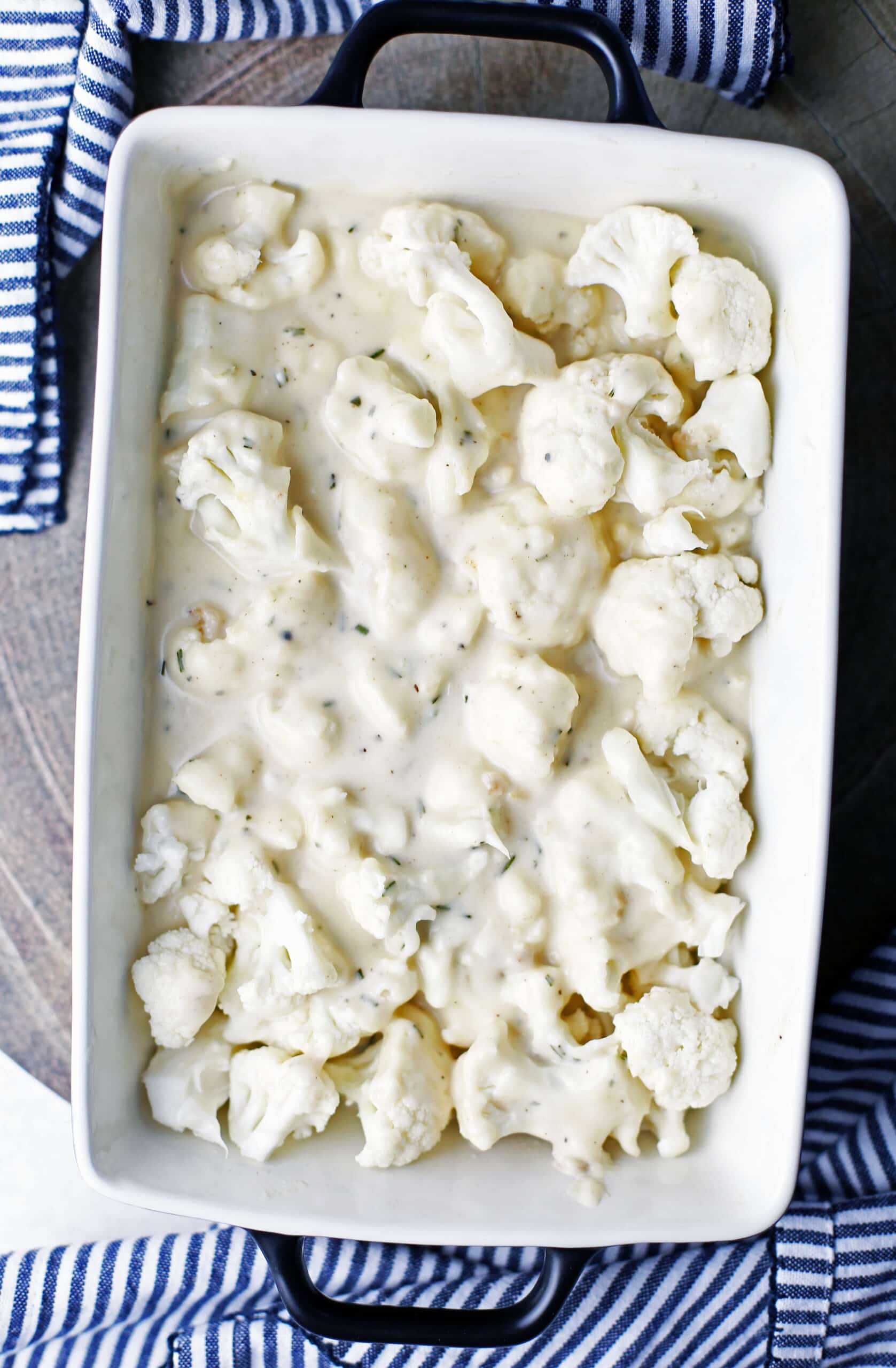 A casserole dish with raw cauliflower florets and cheese white sauce in it.