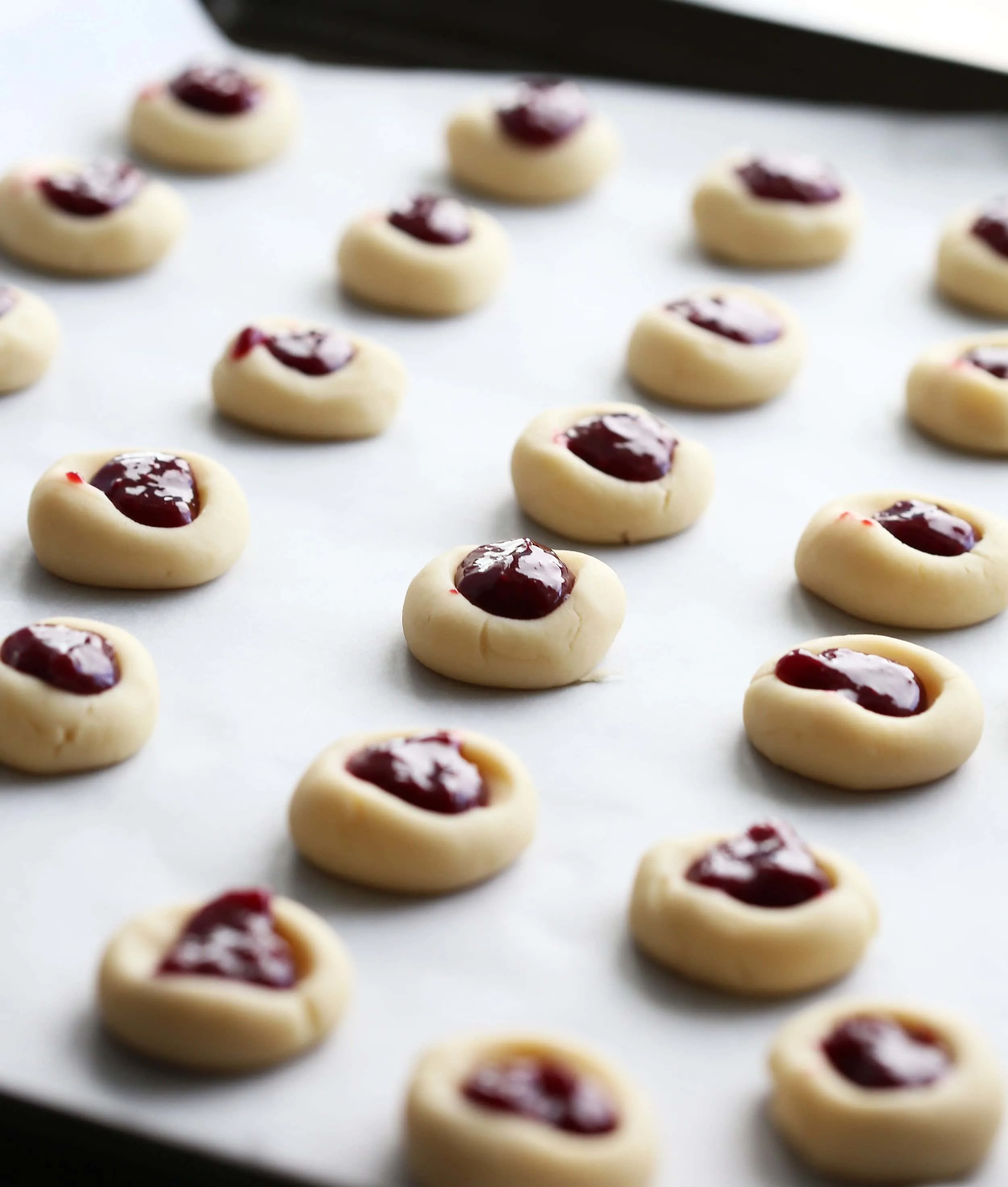 Unbaked shortbread cookies with a thumbprint indentation that’s filled with raspberry jam on a baking sheet.