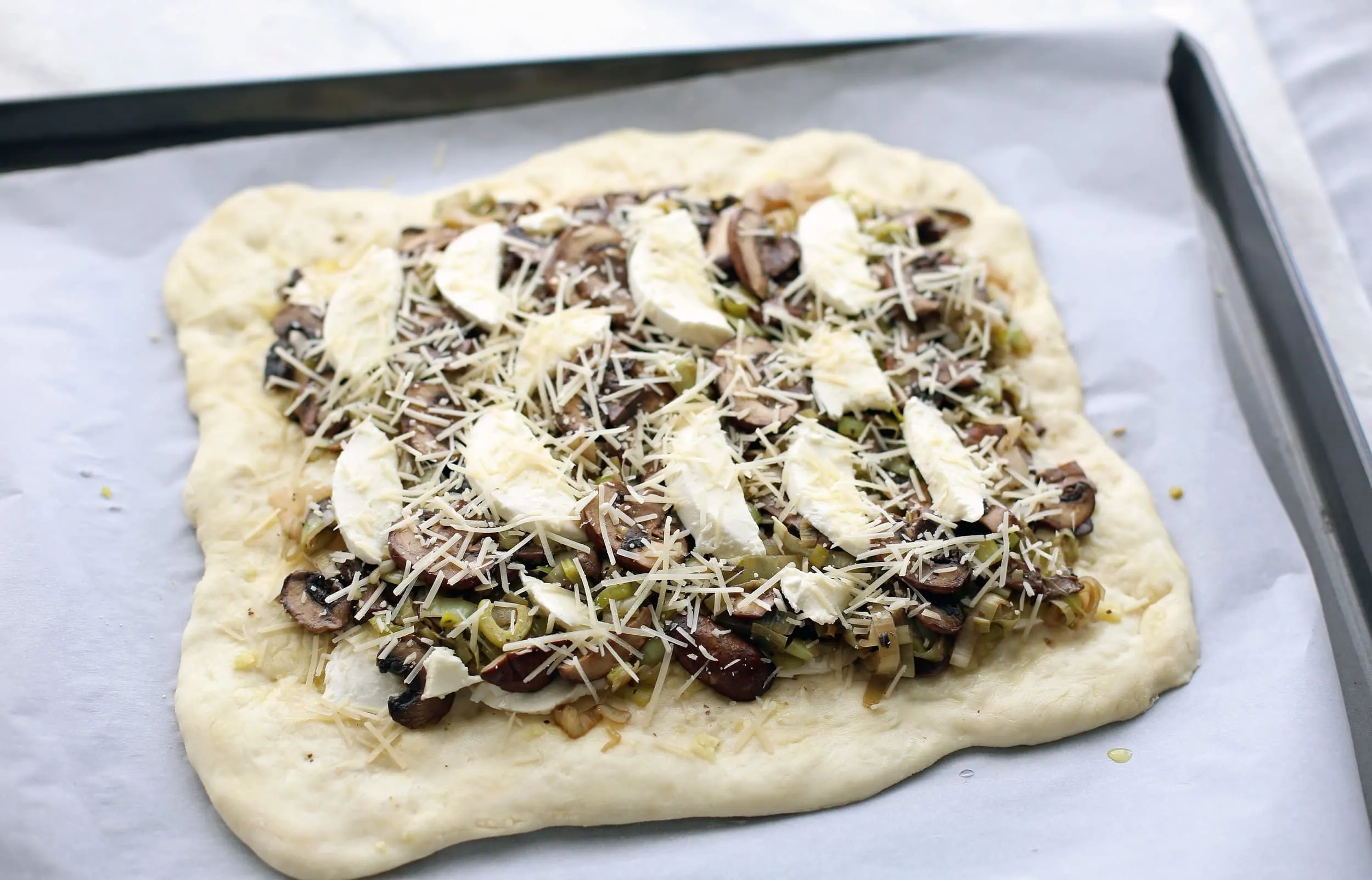 Pizza dough, shaped into a rectangle, with leeks, mushrooms, mozzarella, and parmesan on top.