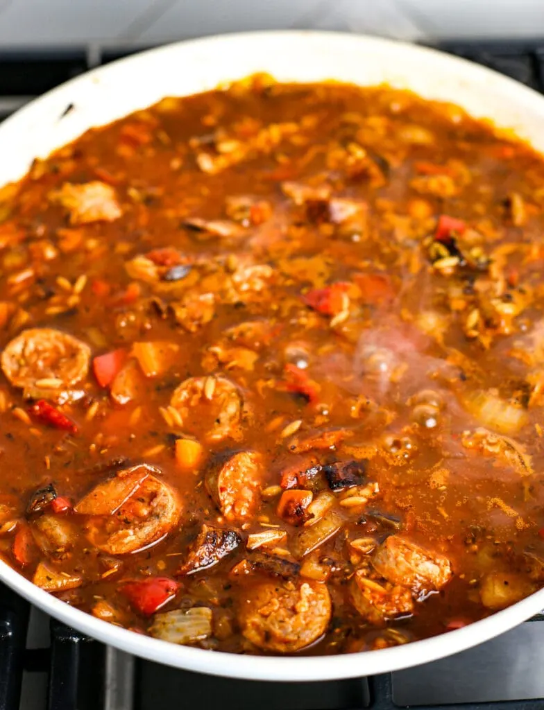 Sliced cooked Italian sausage, sautéed onions and peppers, chicken broth, and marinara sauce in a white pan.