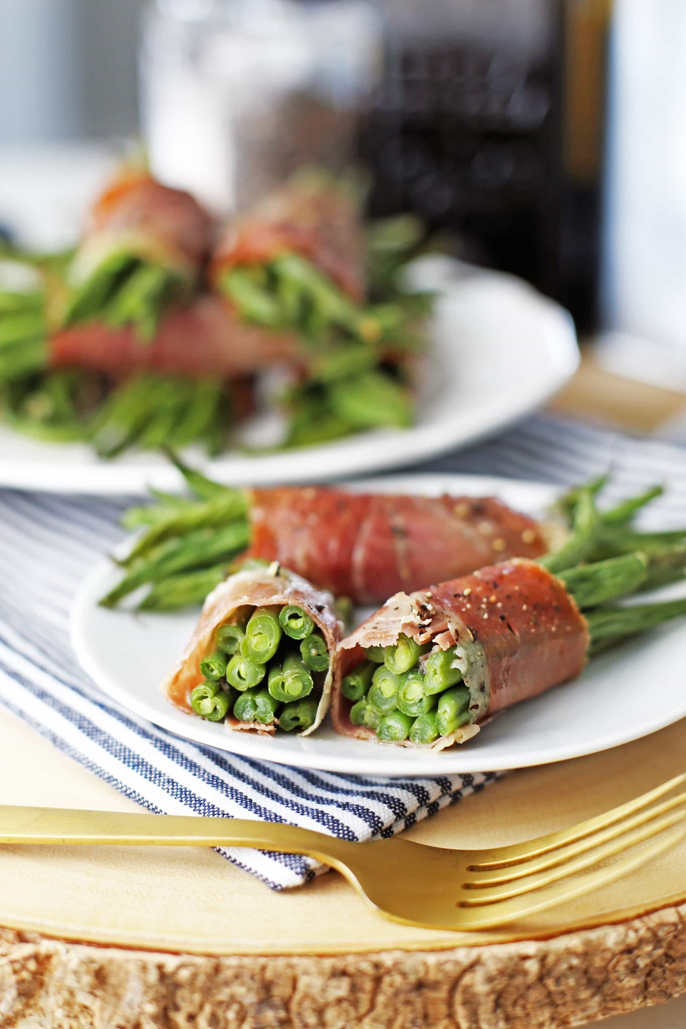 A prosciutto wrapped green bean bundle cut in half on a white plate with more bundles on a plate in the background.
