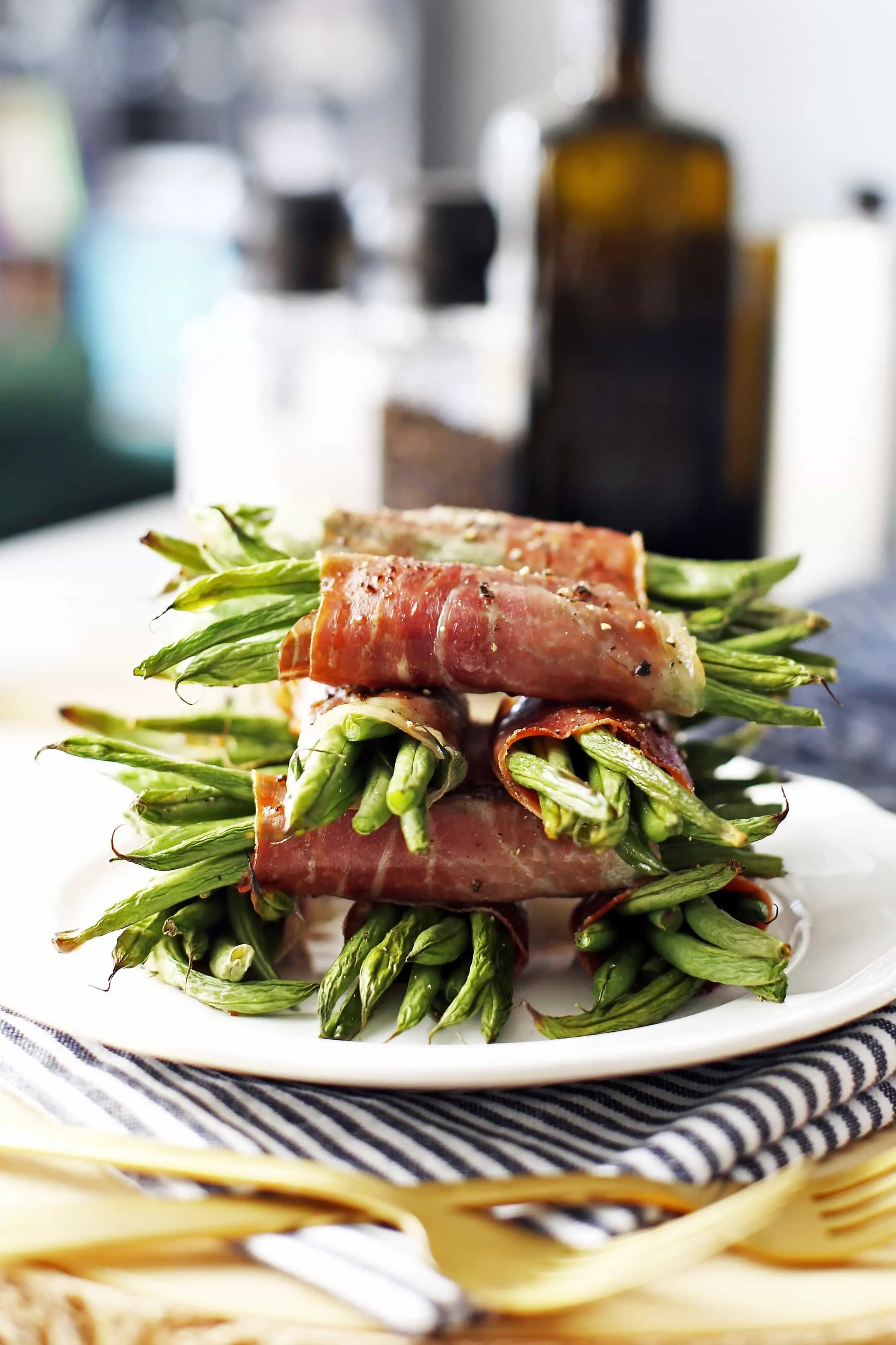 A side view of neatly stacked prosciutto wrapped green bean bundles on a white plate.