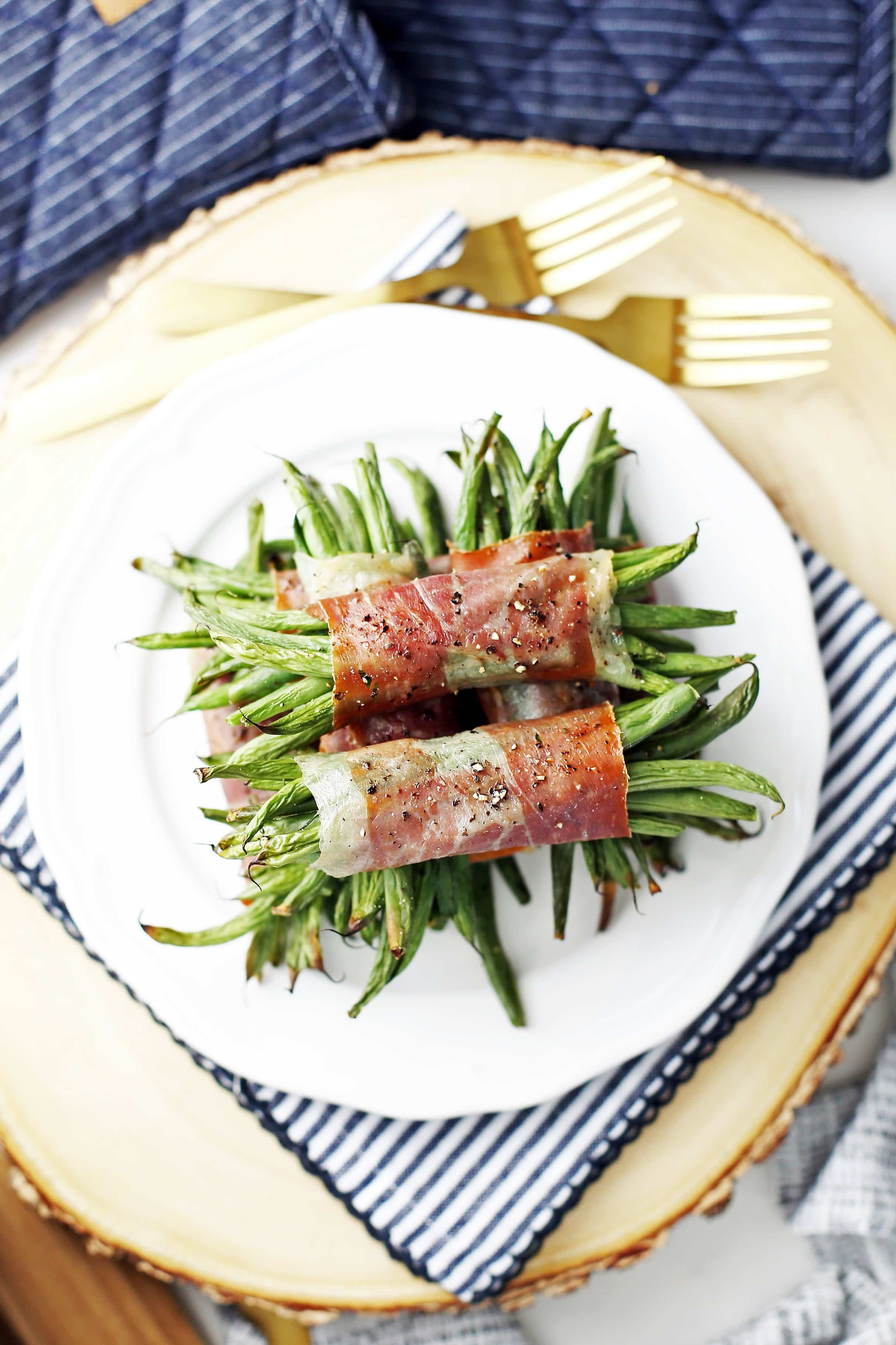Overhead view of prosciutto wrapped green beans piled on a white plate on top of a round wooden board.