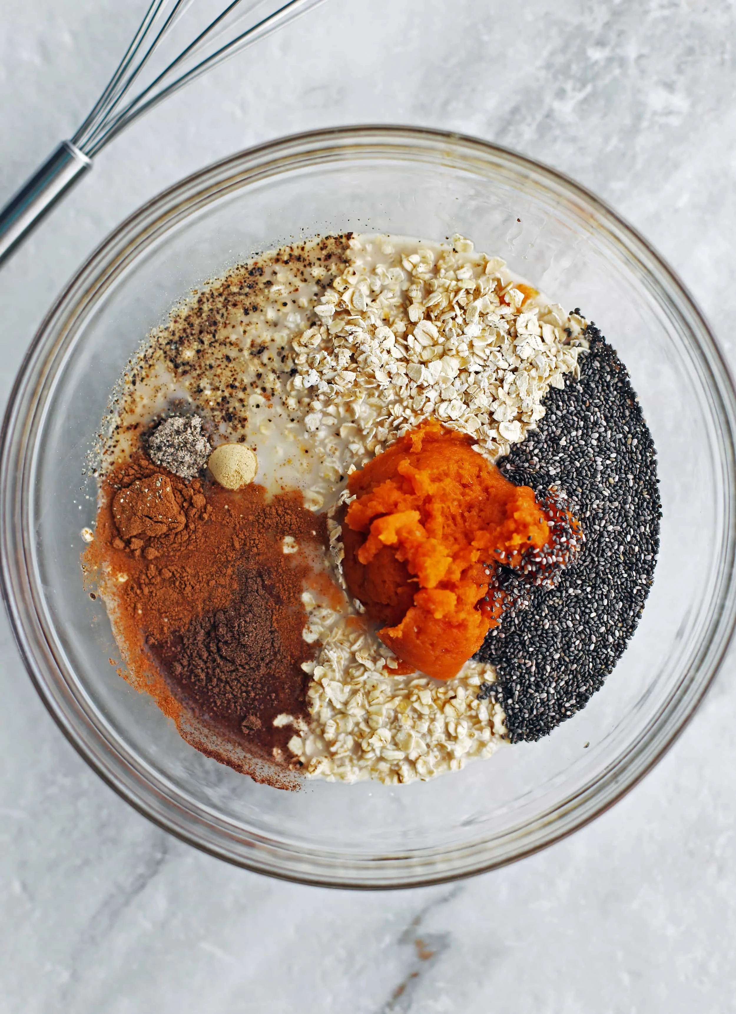 A large glass bowl containing rolled oats, pumpkin puree, chia seeds, spices, almond milk, and maple syrup.