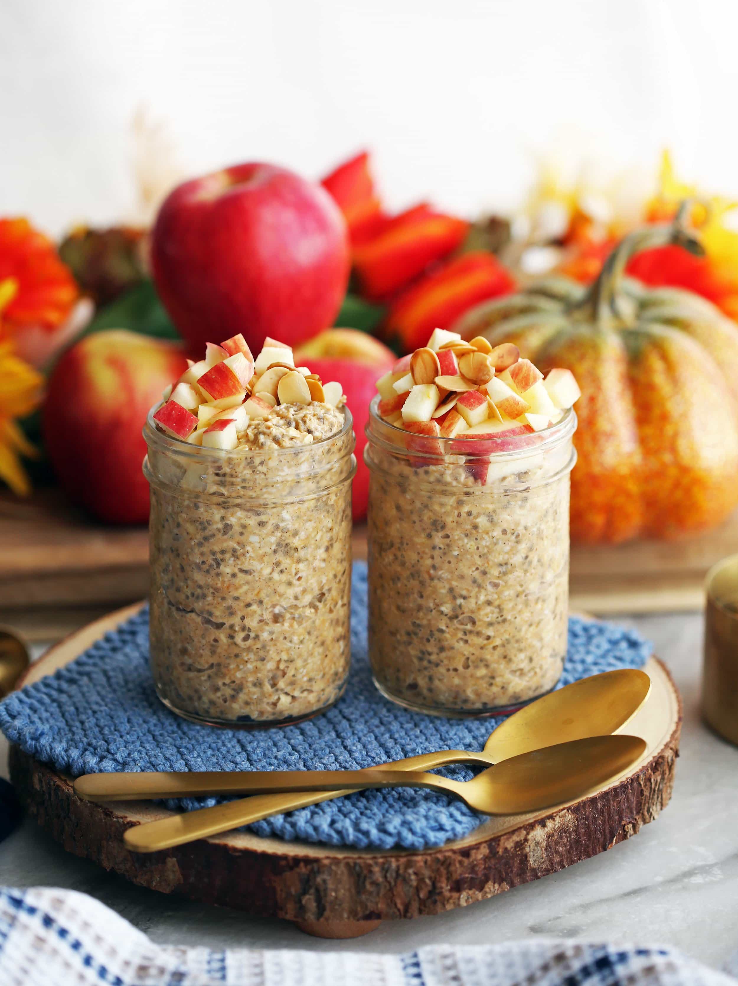 Chai spiced pumpkin overnight oats topped with diced apples in two mason jars on a wooden platter with two spoons.