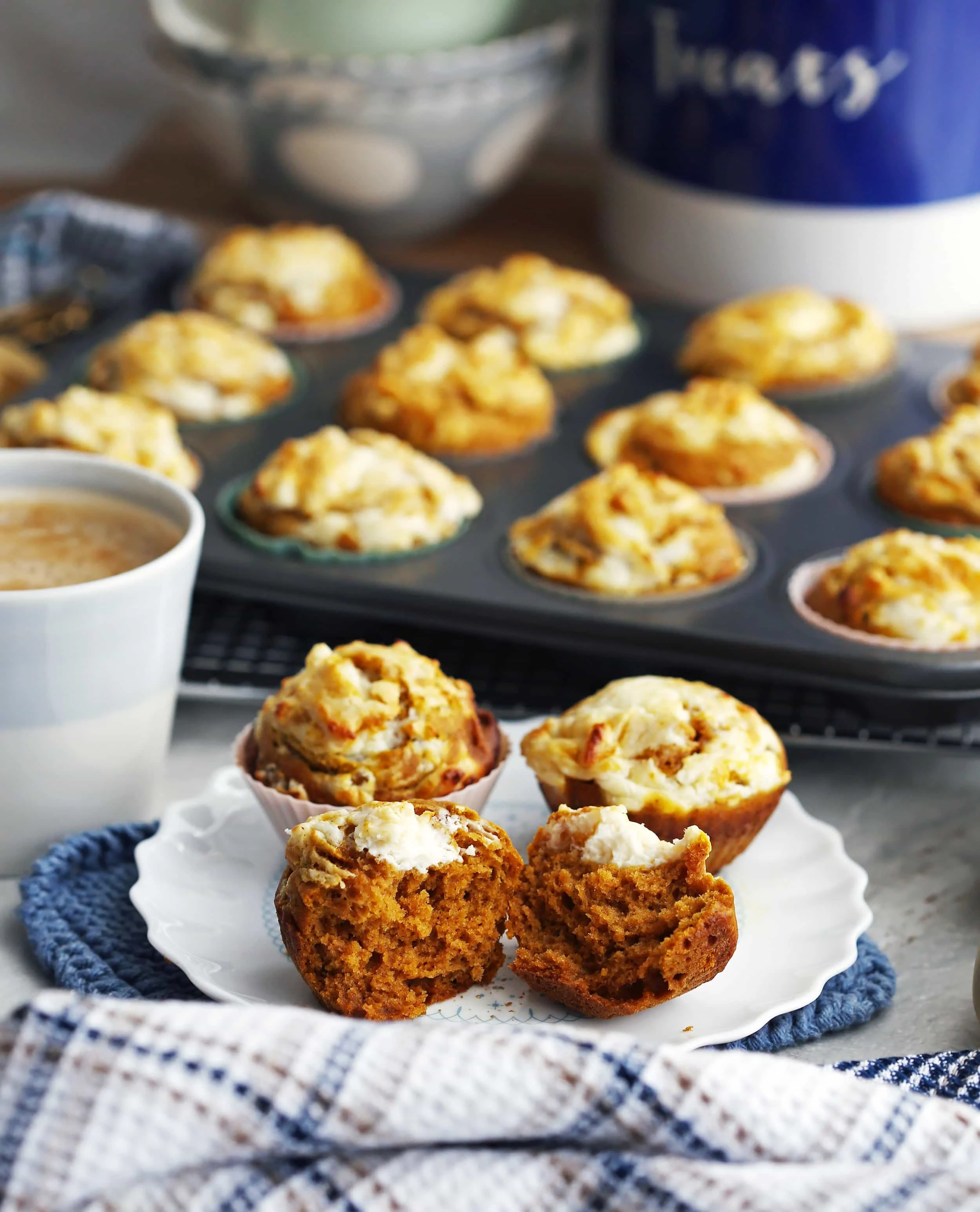 Three pumpkin cream cheese muffins (one muffin pulled in half) on a white plate with more muffins behind them.