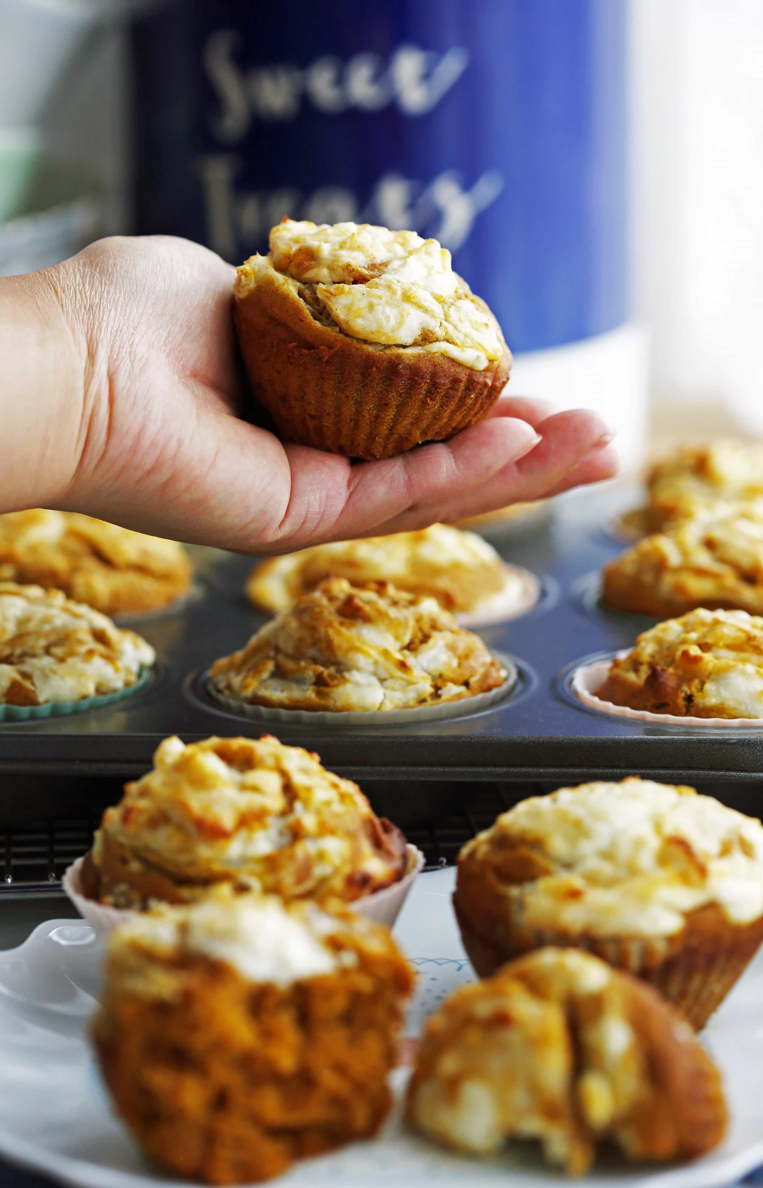A pumpkin cream cheese muffin held in the palm of a hand with more muffins on a plate and muffin pan too.