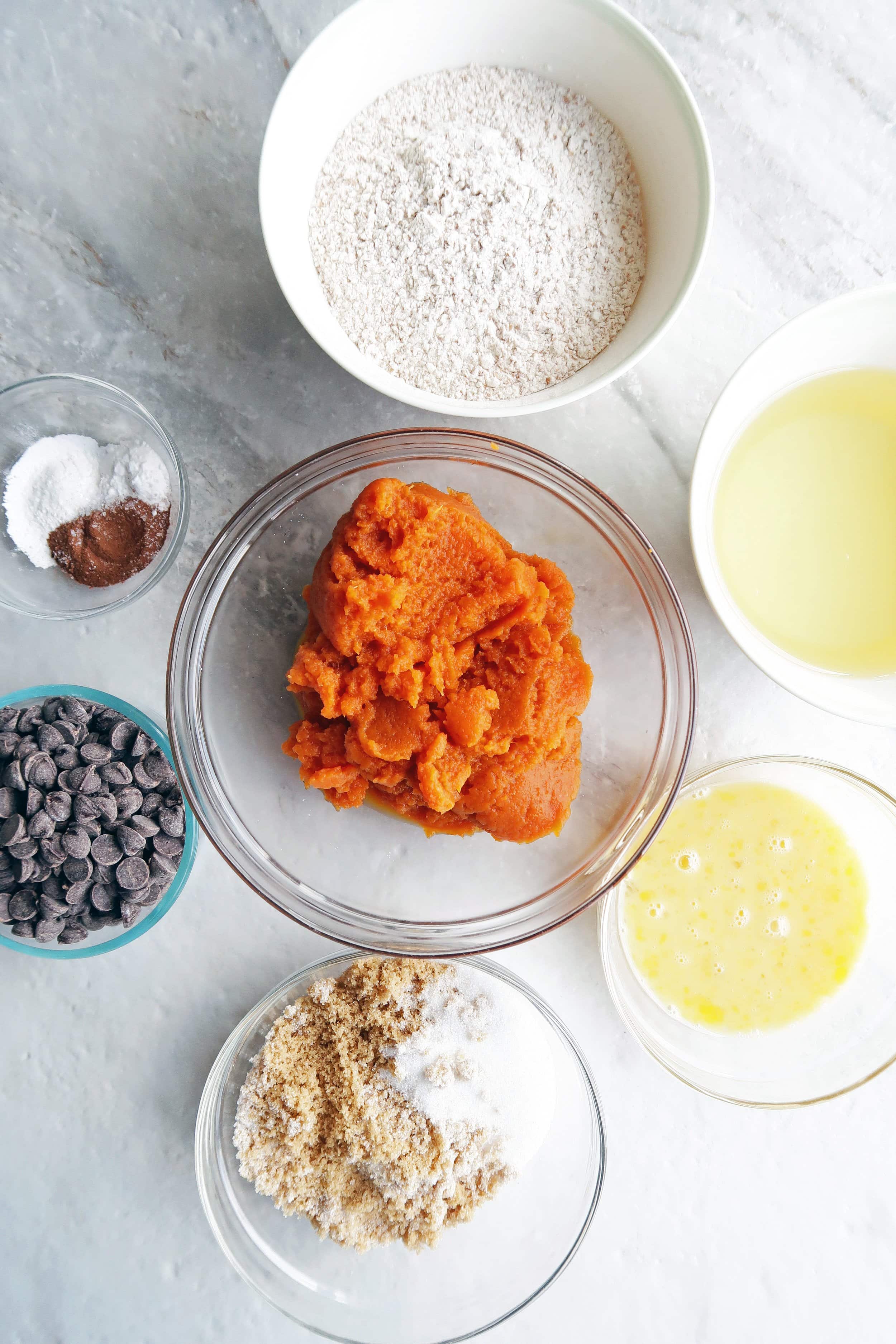 Bowls of chocolate chips, sugar, oil, pumpkin puree, baking soda, baking powder, spices,and eggs.