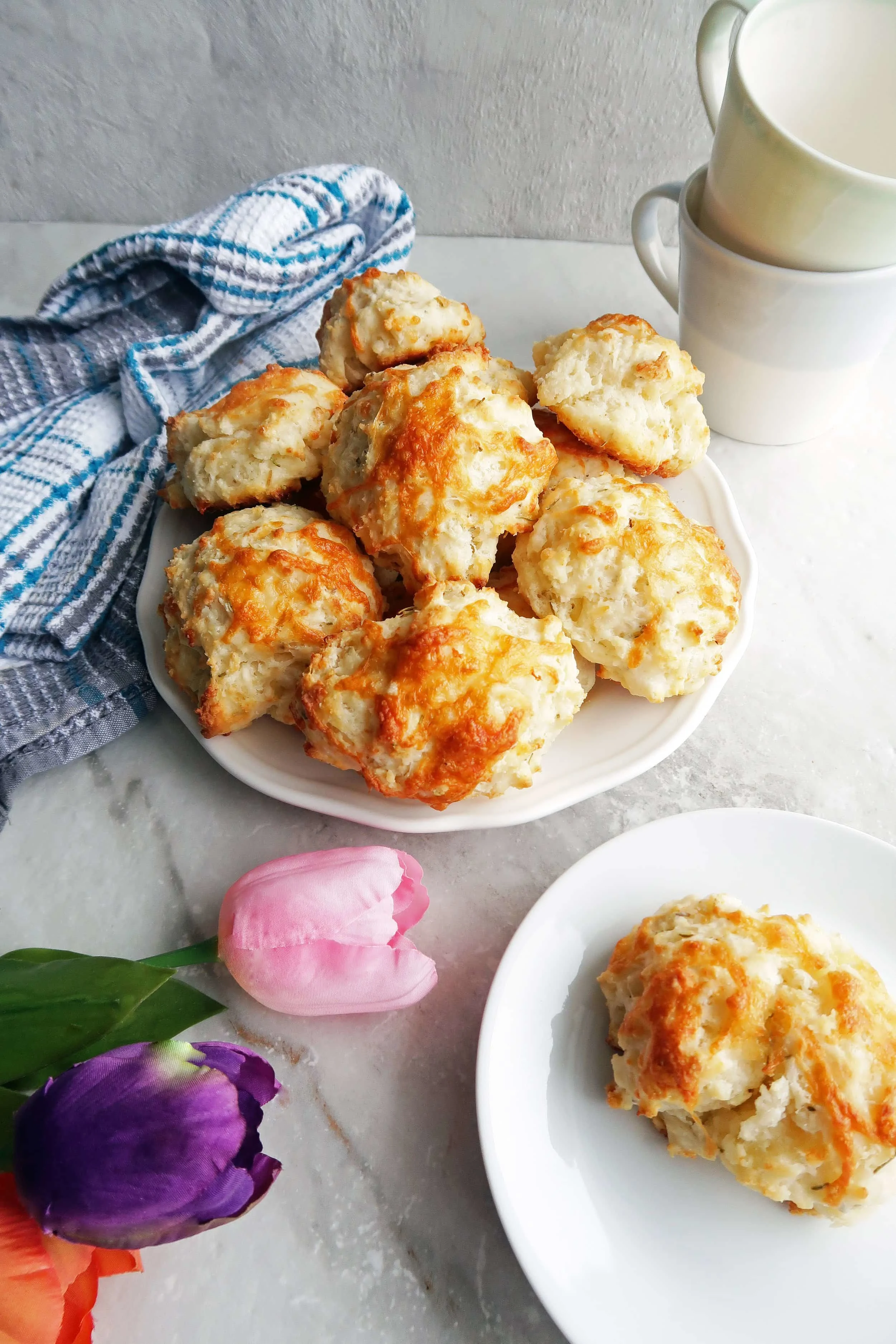 Quick Rosemary Cheddar Drop Biscuits piled on a white plate along with a single biscuit on a plate,tulips, a kitchen towel, and cups.