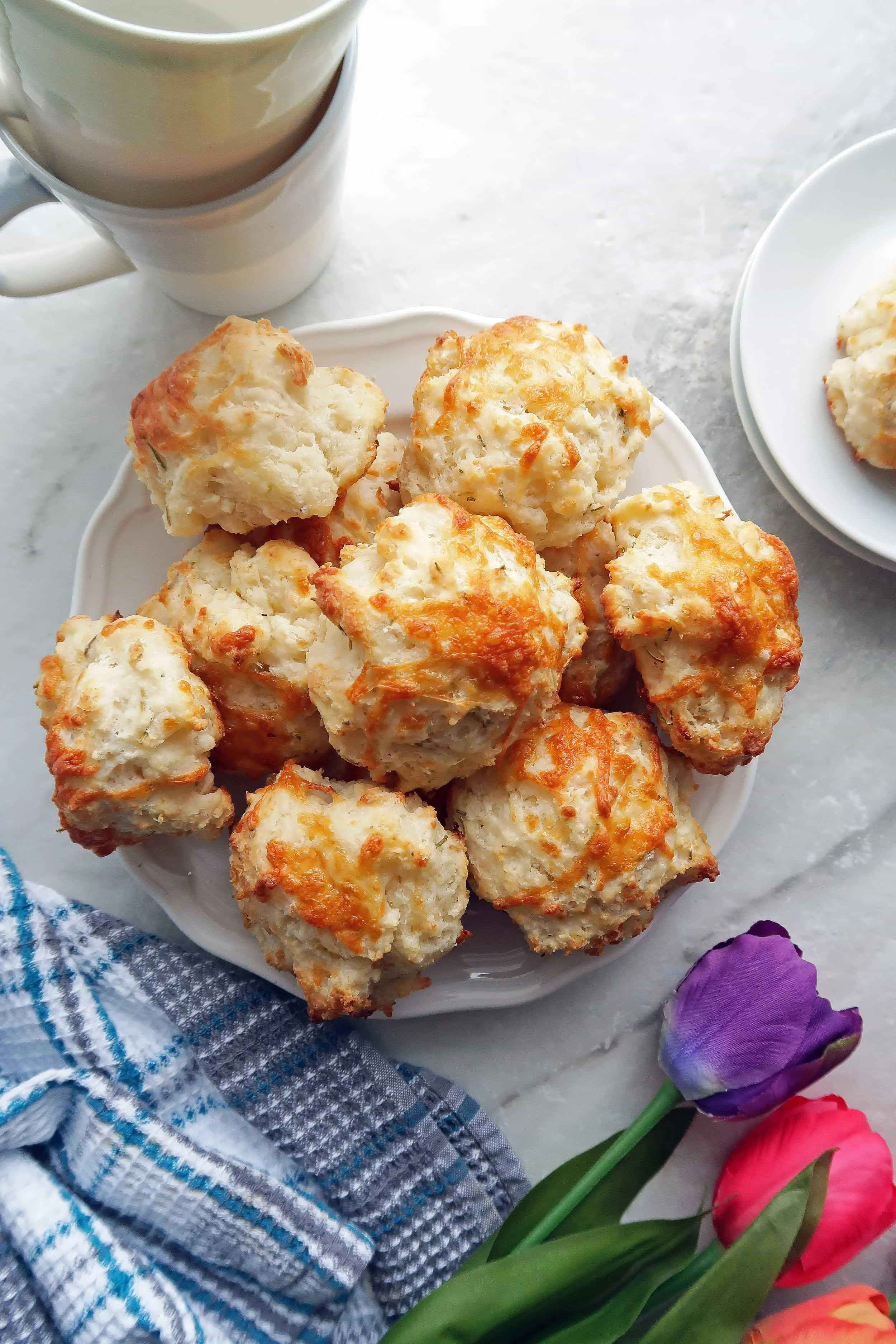 Overhead view of Quick Rosemary Cheddar Drop Biscuits piled on a white plate; tulips, a kitchen towel, cups, and plates around them.