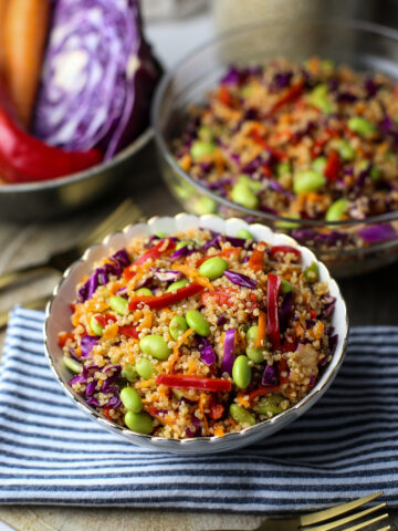 A small bowl of quinoa edamame salad with peppers, cabbage, and carrots with more salad in a glass bowl behind it.