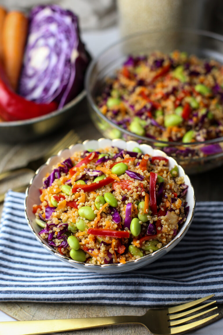 A small bowl of quinoa edamame salad with peppers, cabbage, and carrots with more salad in a glass bowl behind it.