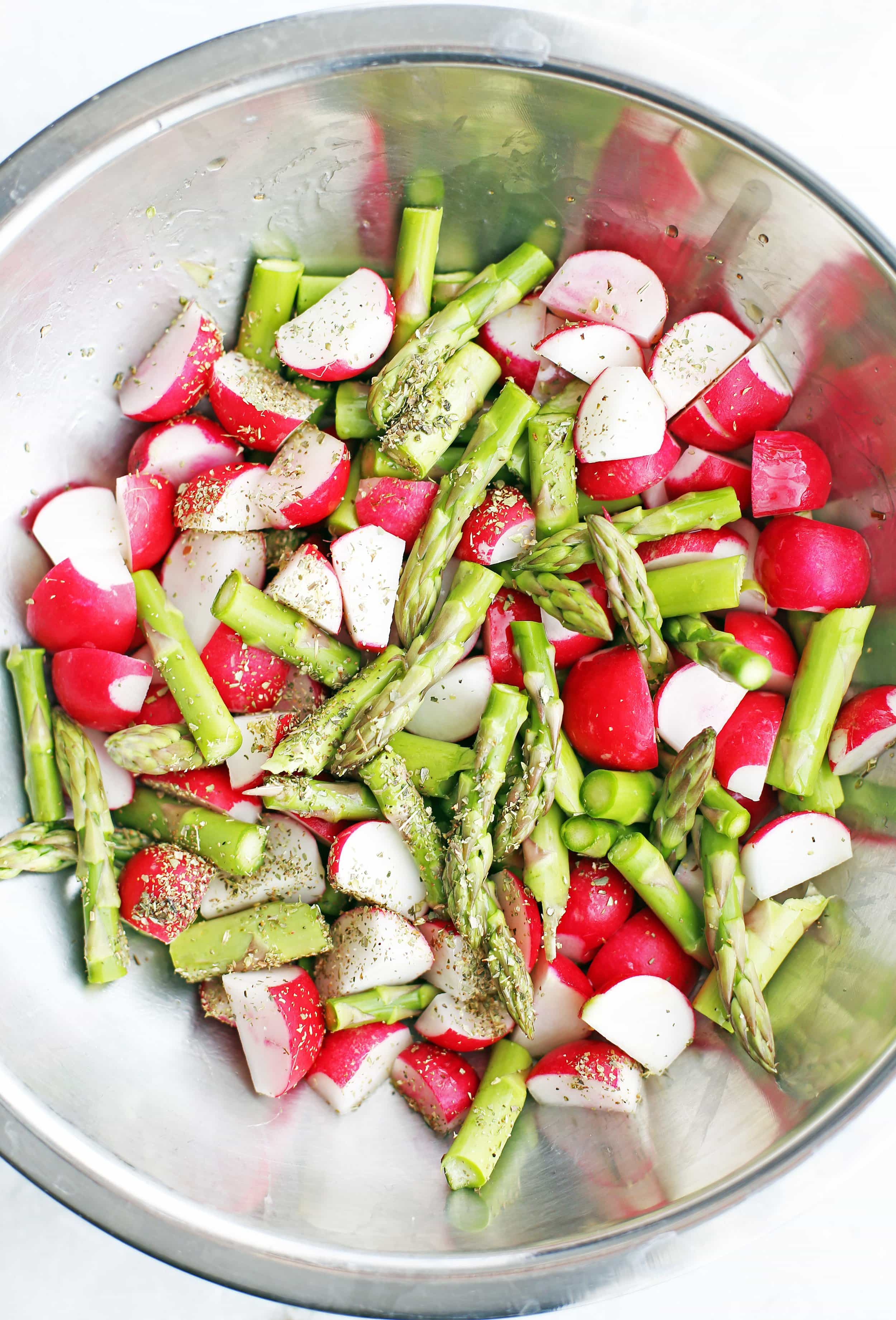 Chopped asparagus and radishes, olive oil, salt, pepper, and dried oregano in a metal bowl.