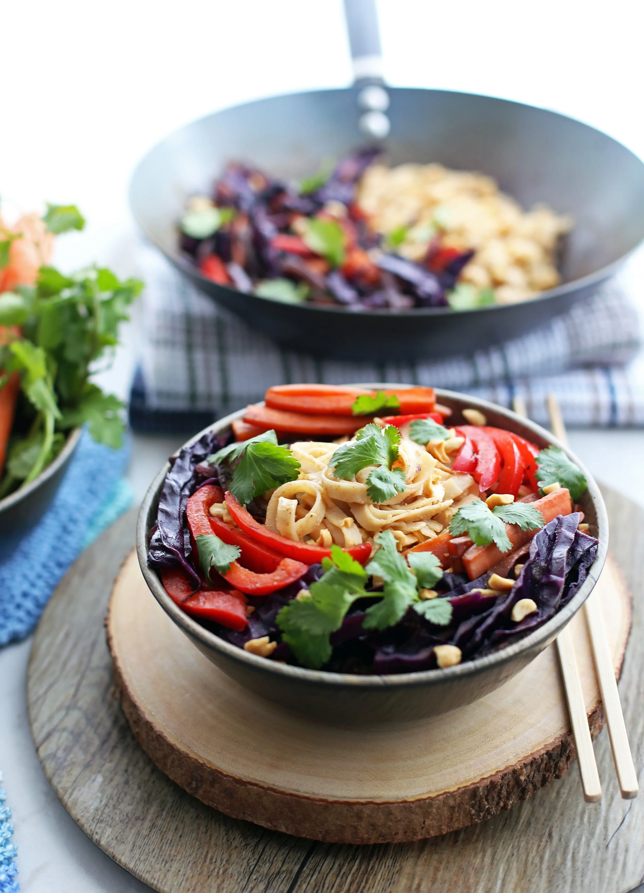 Stir-Fry Vegetables and Noodles with Peanut Sauce in a wooden bowl and in a wok behind it.