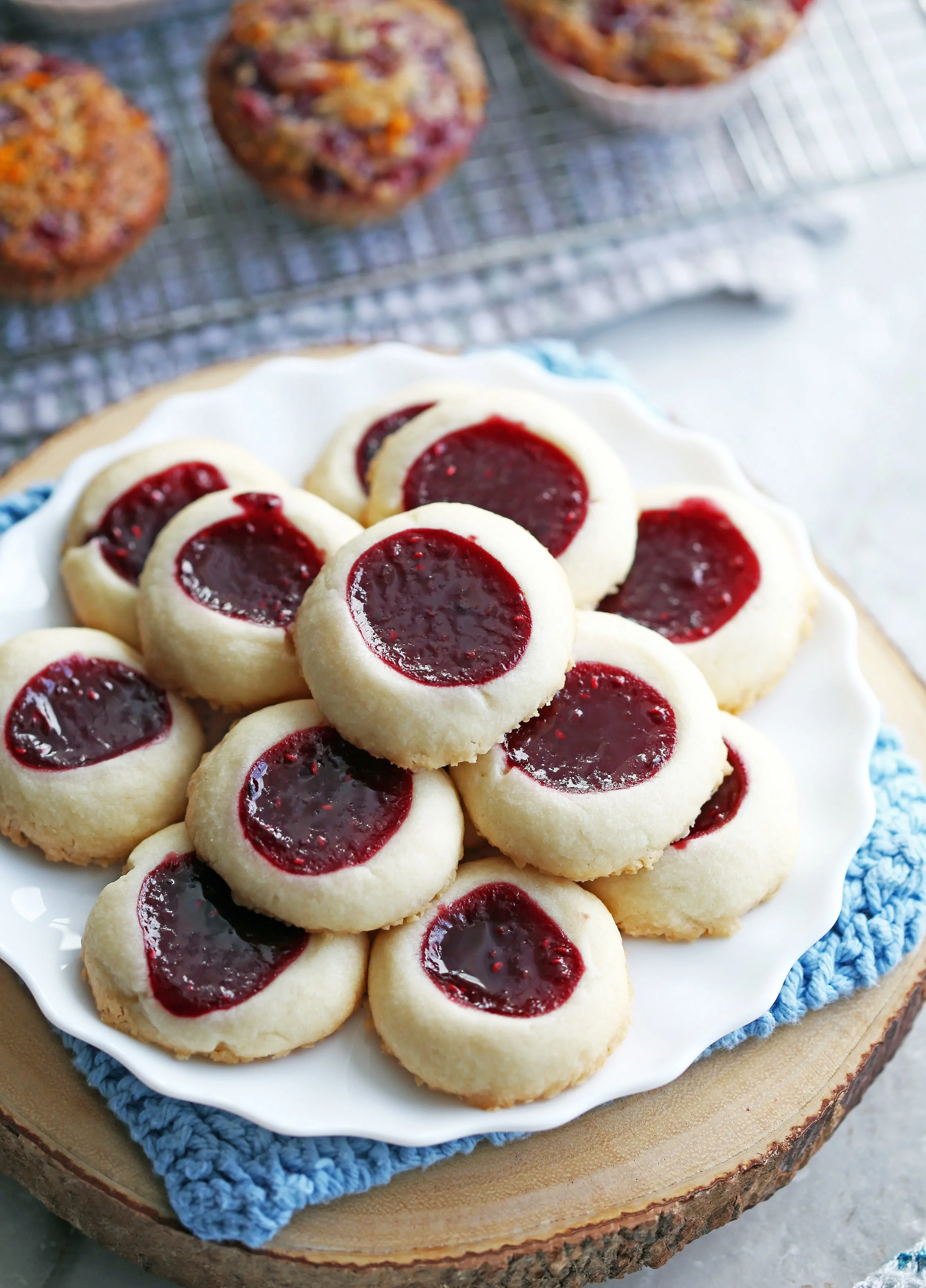 Raspberry shortbread thumbprint cookies piled on a white plate that’s on top of a wooden platter.