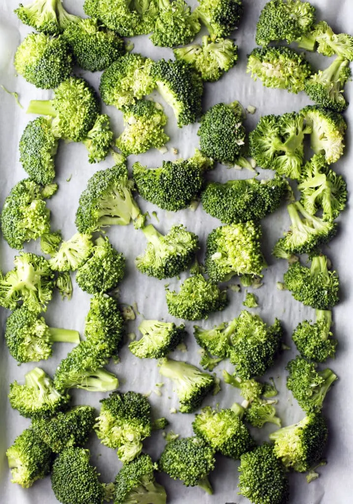 Raw broccoli florets and minced garlic tossed in olive oil on parchment paper lined baking sheet.