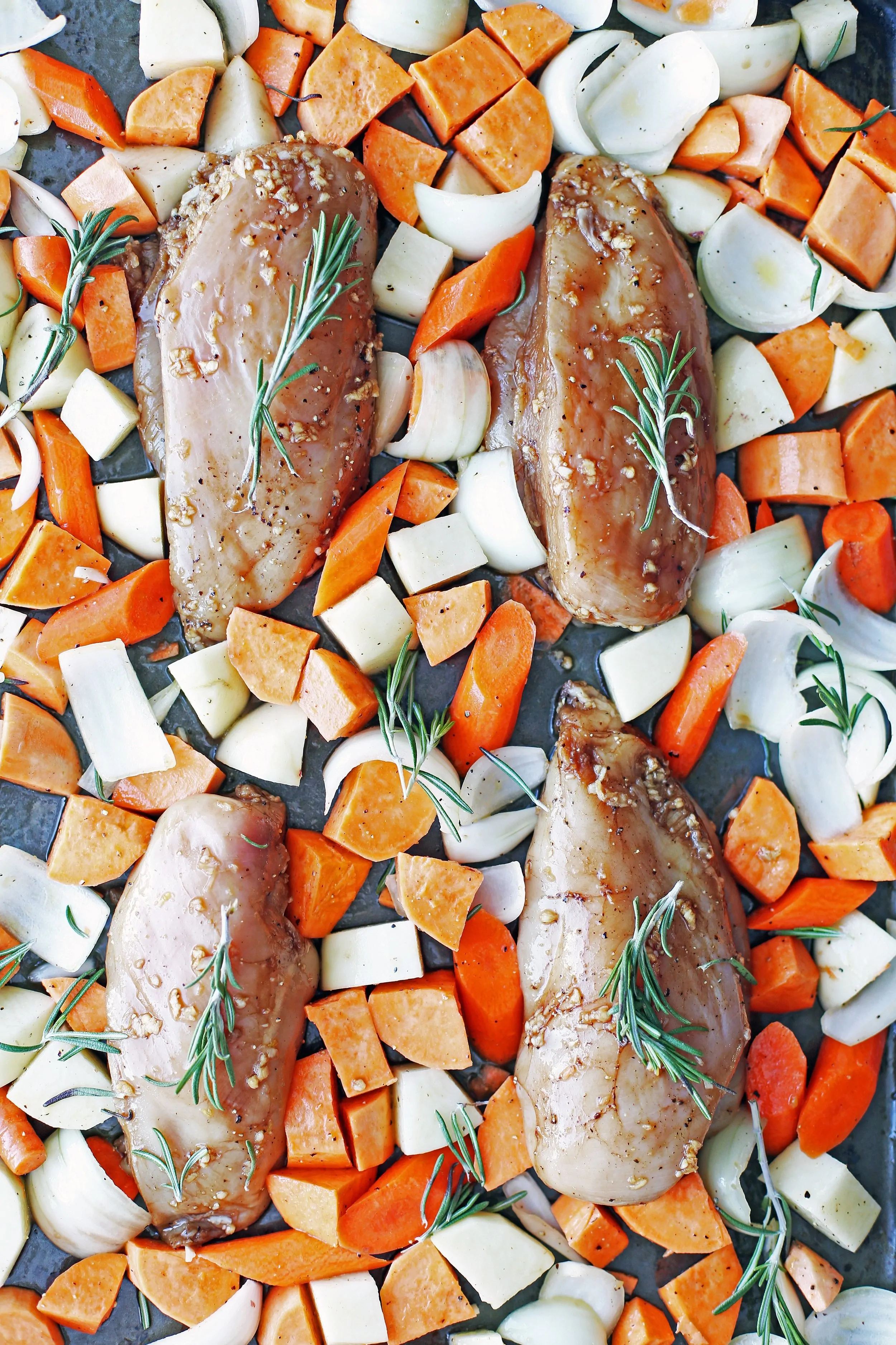 Four marinated chicken breasts, chopped carrots, potatoes, onions, and rosemary sprigs placed on a baking sheet.