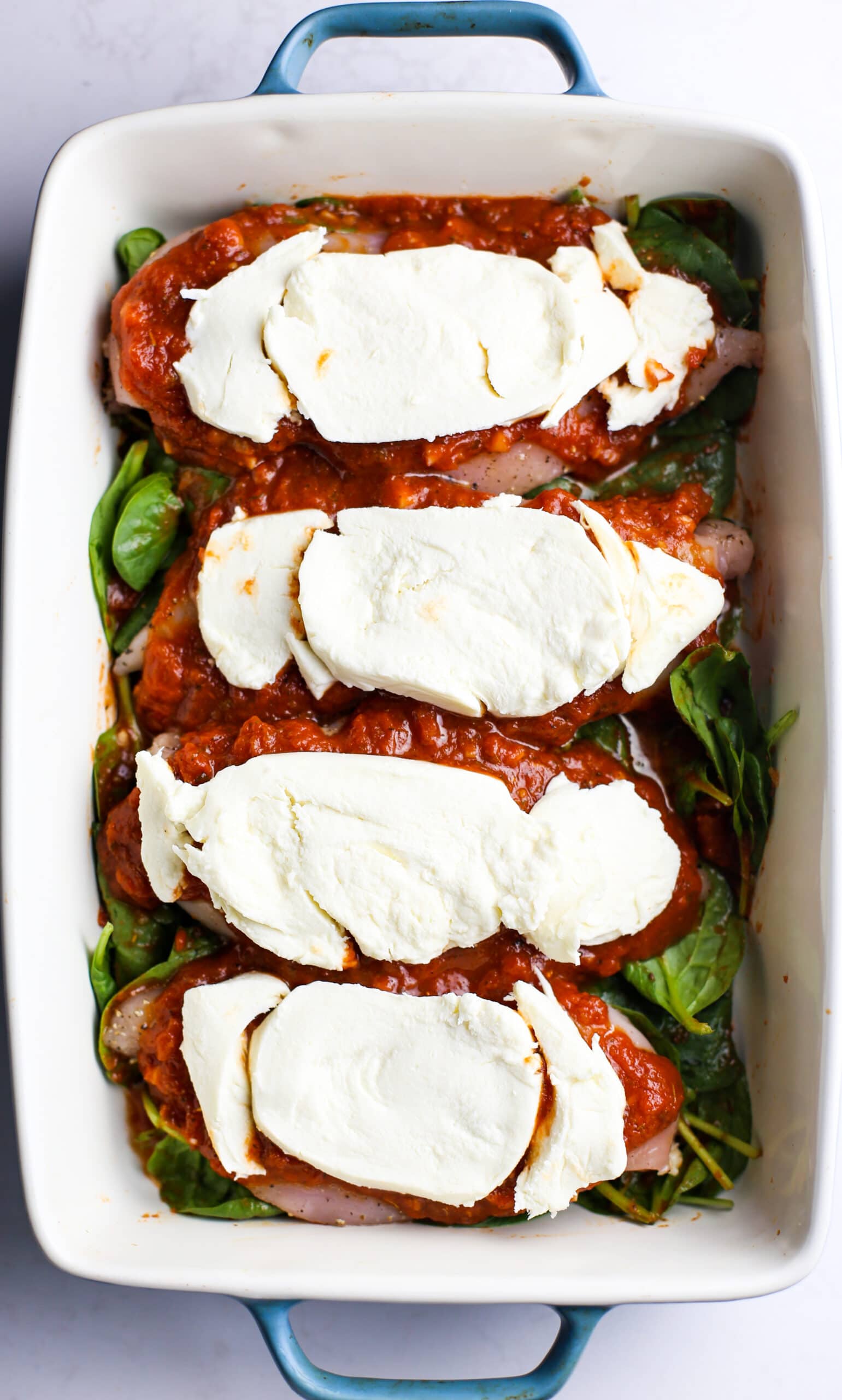Four raw chicken breasts topped with marinara sauce and mozzerella cheese on a bed of spinach in a casserole dish.