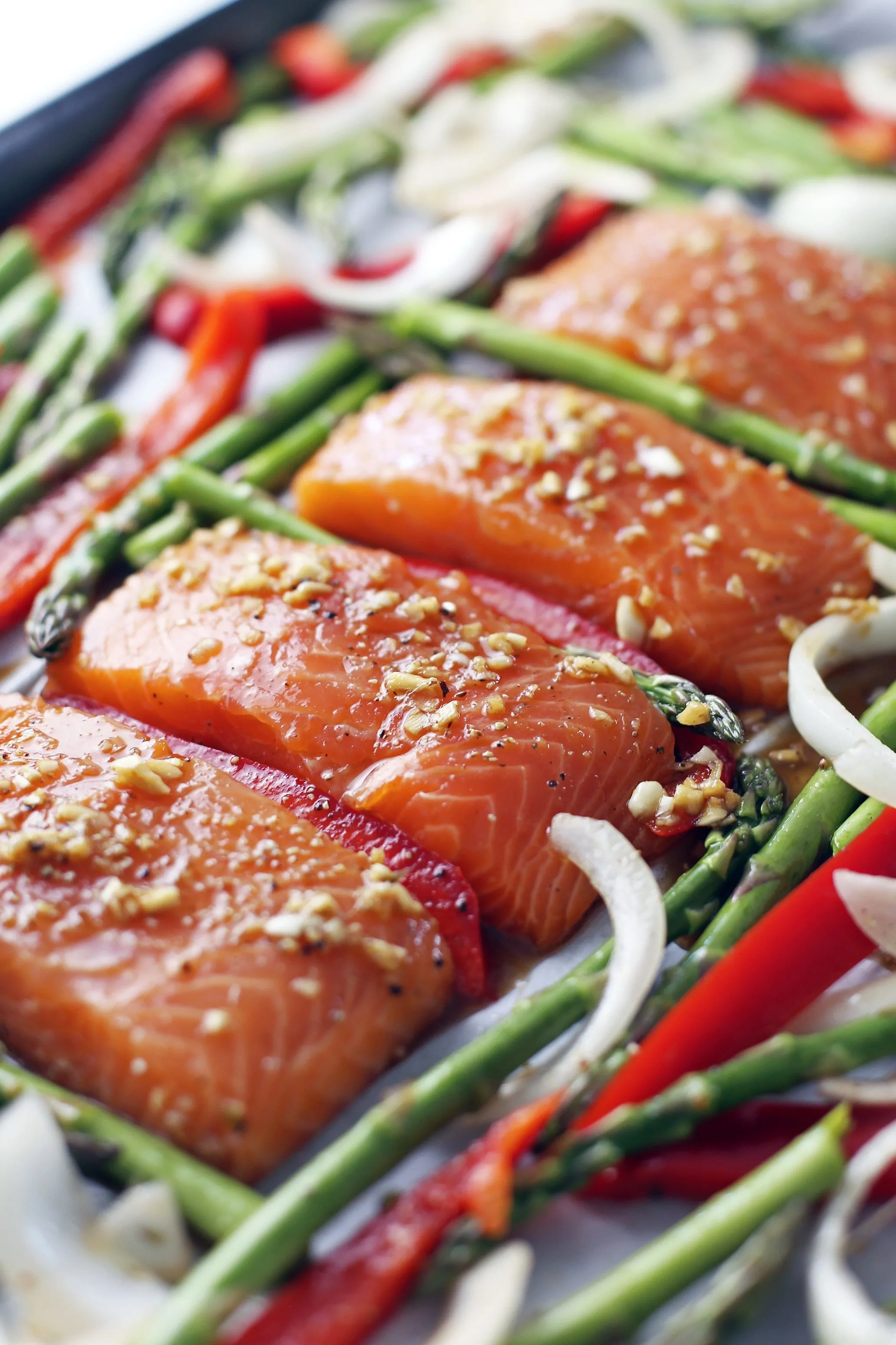 Four raw marinated salmon fillets in a row with asparagus, bell pepper, and onions surrounding them on a large baking sheet.