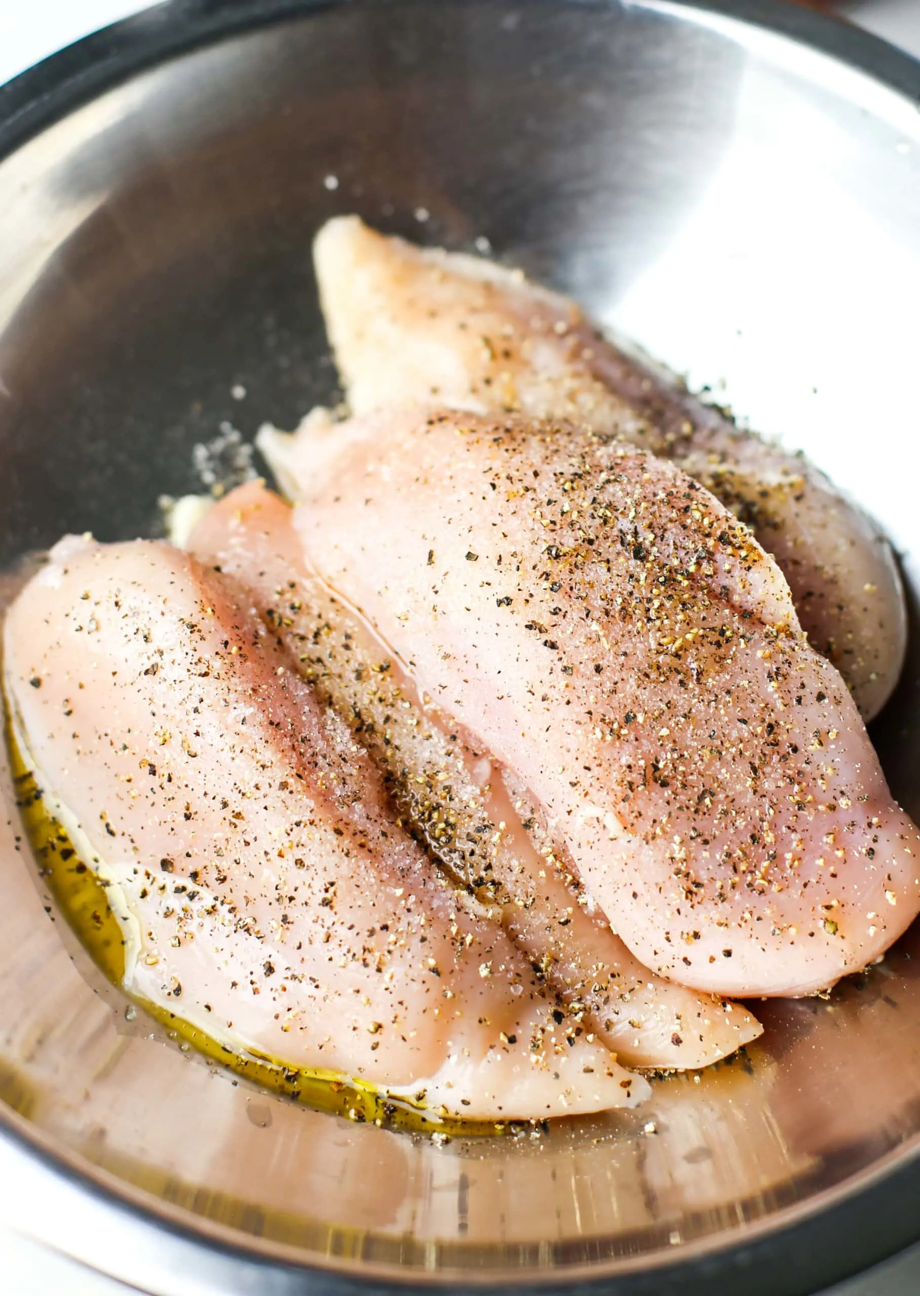 Four raw seasoned chicken breasts in a stainless steel bowl.