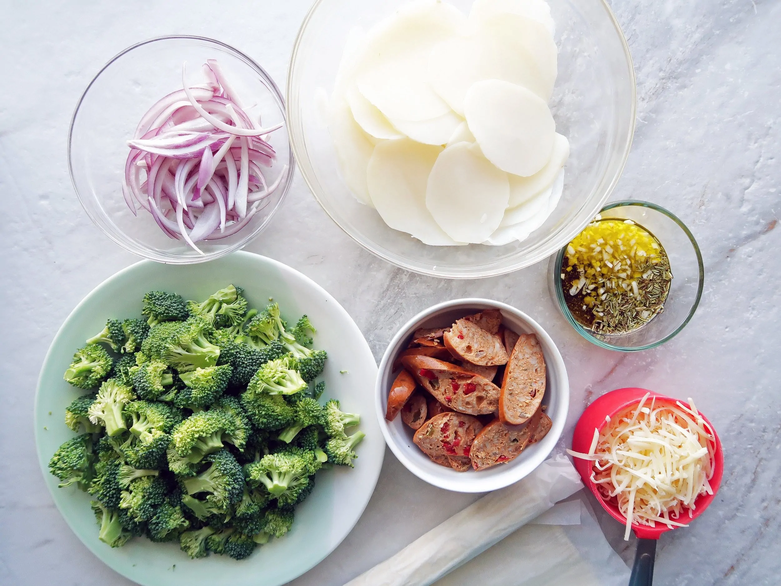 Bowls of broccoli, sliced potatoes, sausage, red onions, and grated cheese.