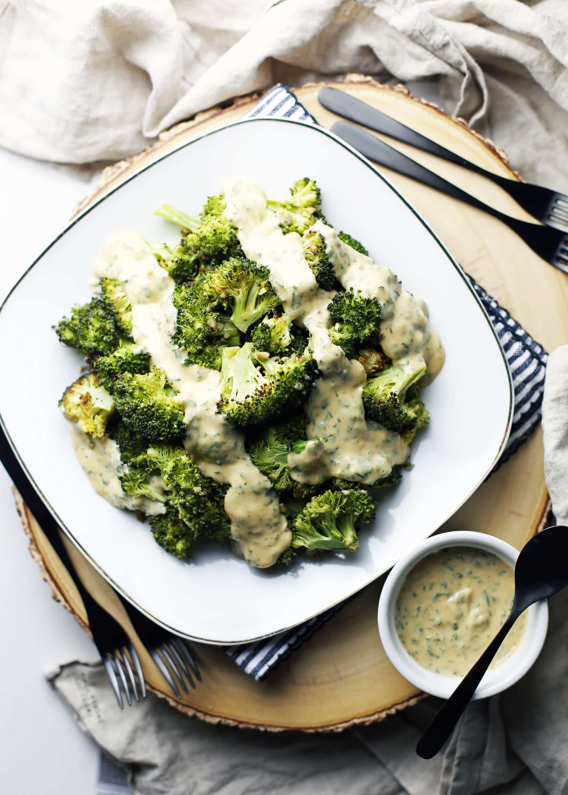 Overhead view of roasted broccoli with parsley lemon hollandaise sauce on a white plate with more sauce in a small ramekin beside it.