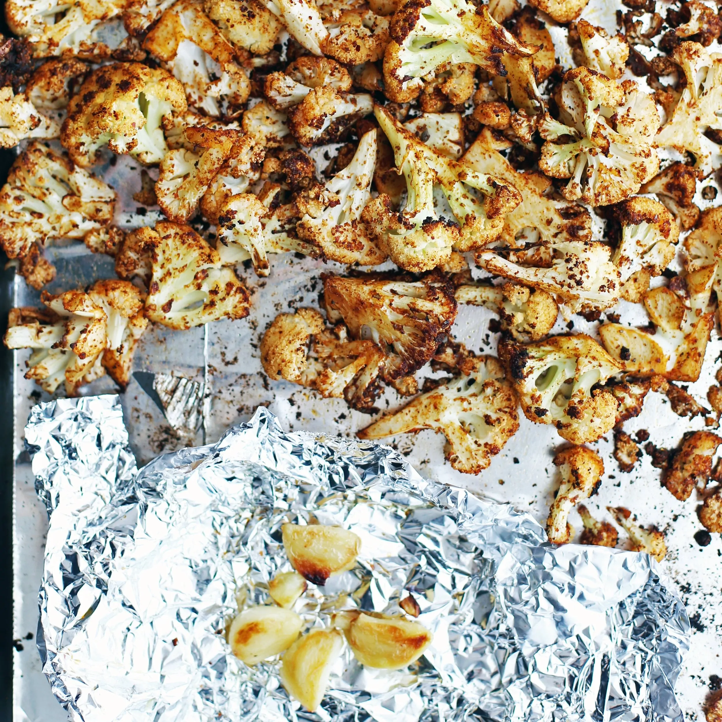 Roasted spiced cauliflower florets and roasted garlic cloves on a baking sheet.