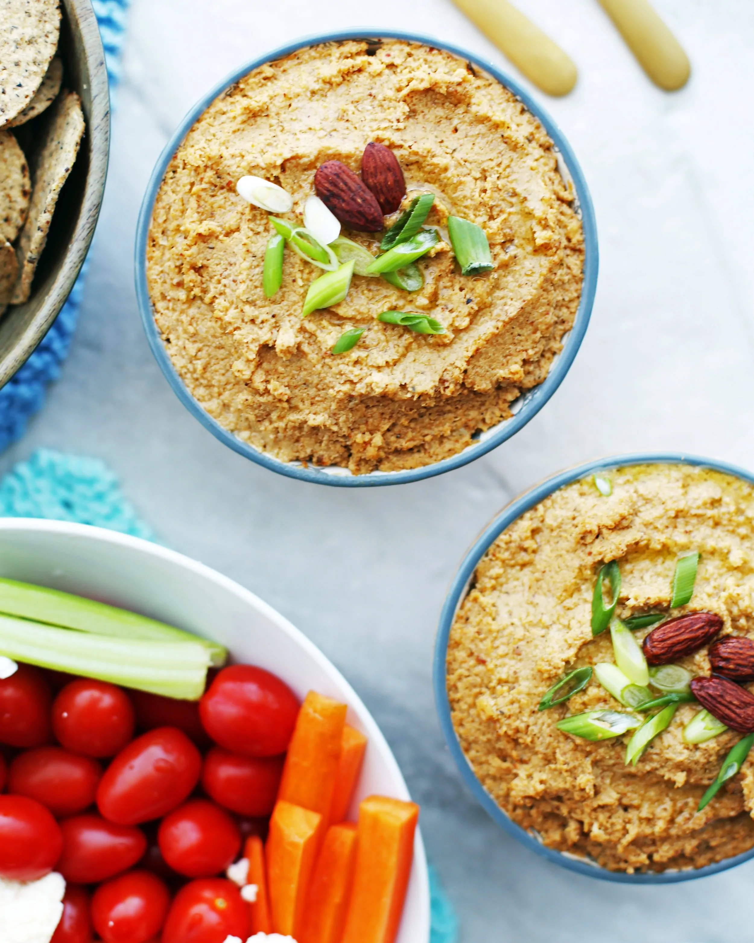 Overhead view of two bowls of spicy roasted cauliflower garlic dip topped with almonds and green onions along with a bowl of chopped vegetables.