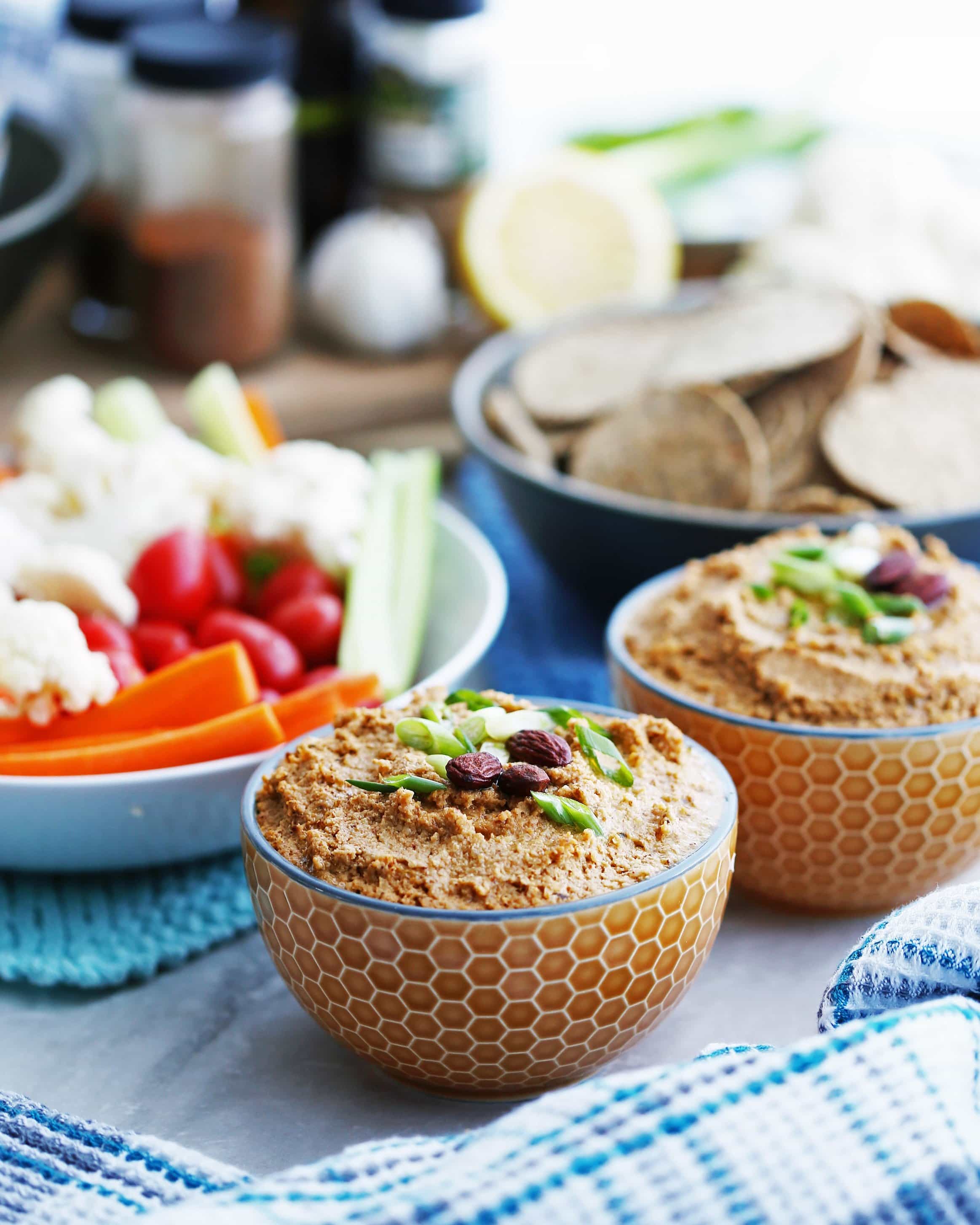 A bowl of spicy roasted cauliflower garlic dip topped with almonds and green onions; another bowl of dip, tortillas, and chopped vegetables in a bowl behind it.