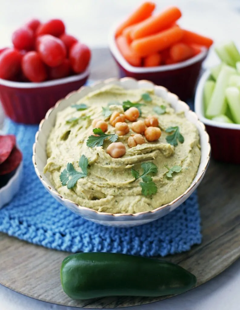 A close angled view of a bowl of jalapeno cilantro hummus with carrots, tomatoes, celery, and crackers.