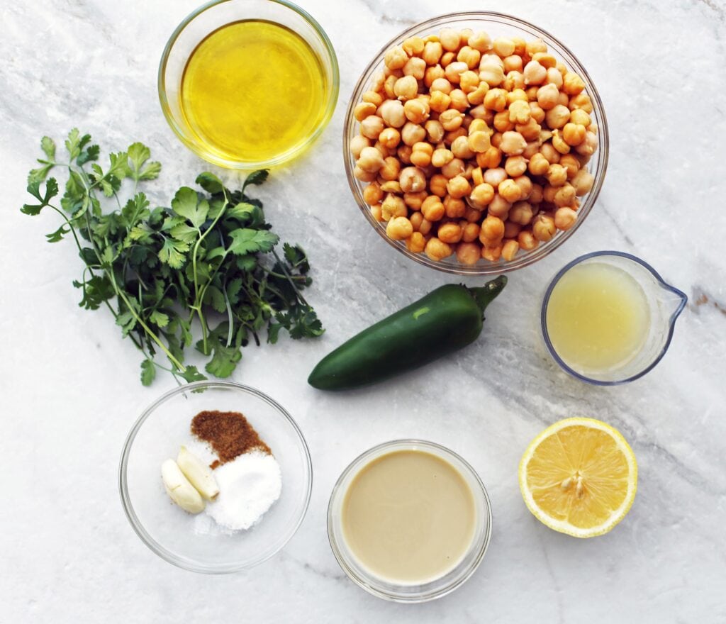 An overhead view of cooked chickpeas, cilantro, jalapeno pepper, tahini, lemon, garlic, olive oil,cumin, and salt.